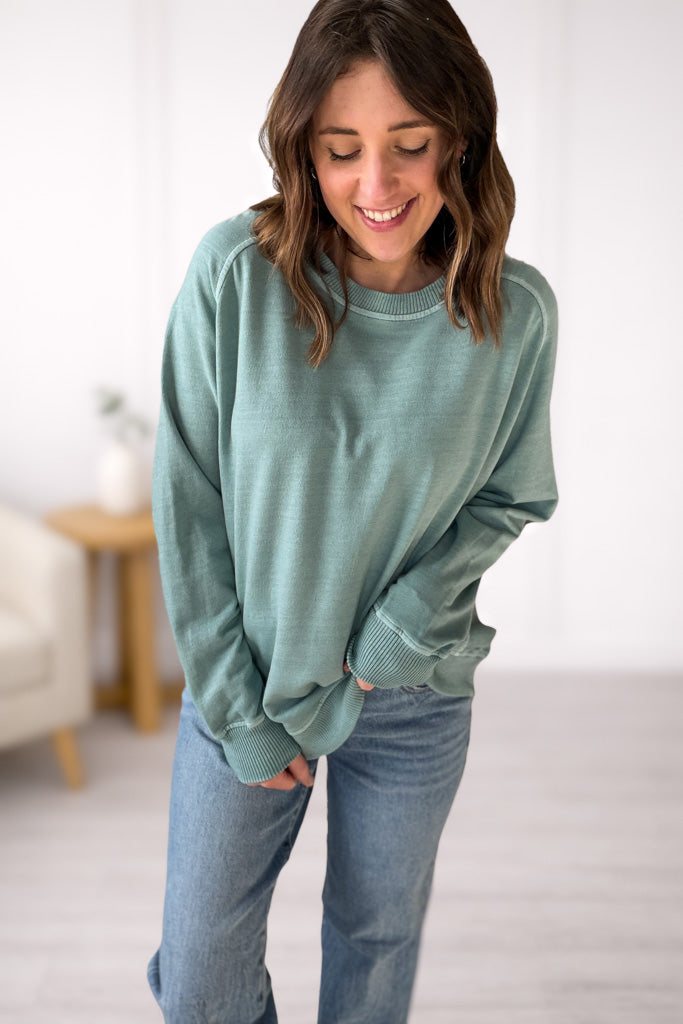 Woman wearing a green/blue sweater and blue jeans indoors.