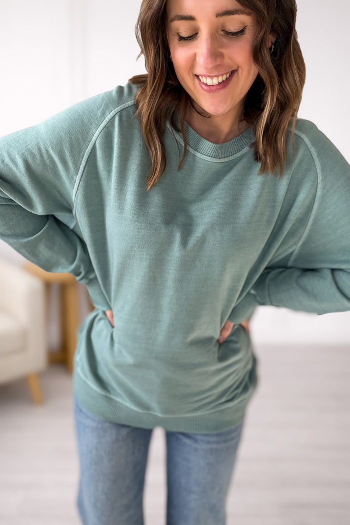 Woman wearing a green/blue sweater and blue jeans indoors.