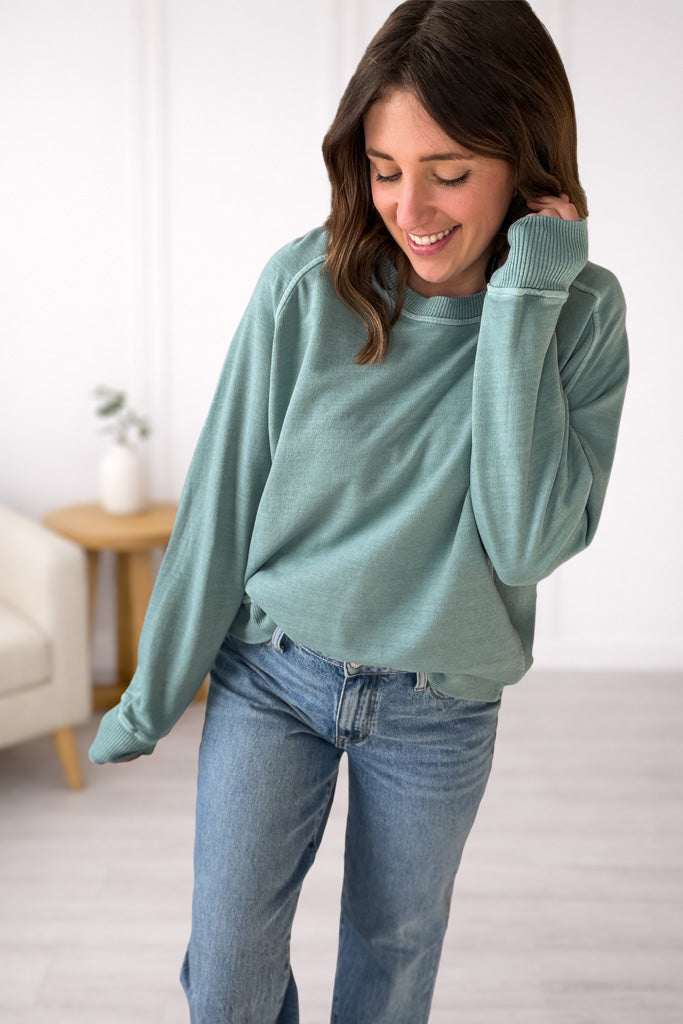 Woman wearing a teal sweater and blue jeans in a room with a white wall and a small table with a plant.