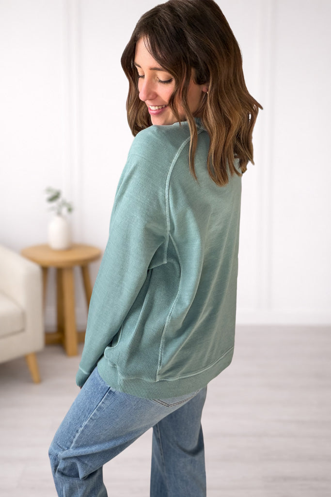Woman wearing a light green sweater and blue jeans in a room with a white wall and wooden table.