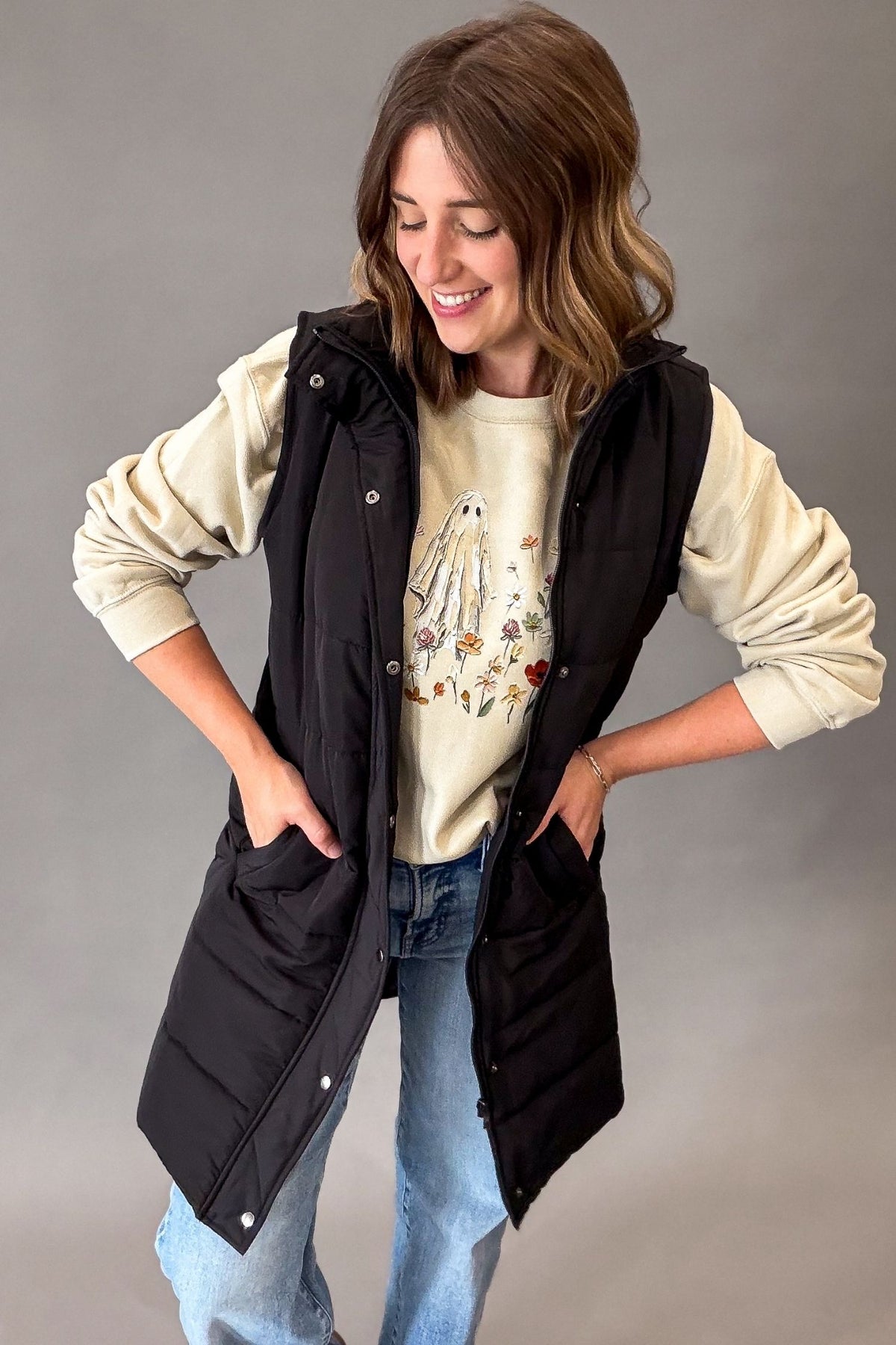 A woman modeling a black sleeveless puffer vest over a cream graphic sweatshirt and blue jeans, standing against a neutral gray studio background.