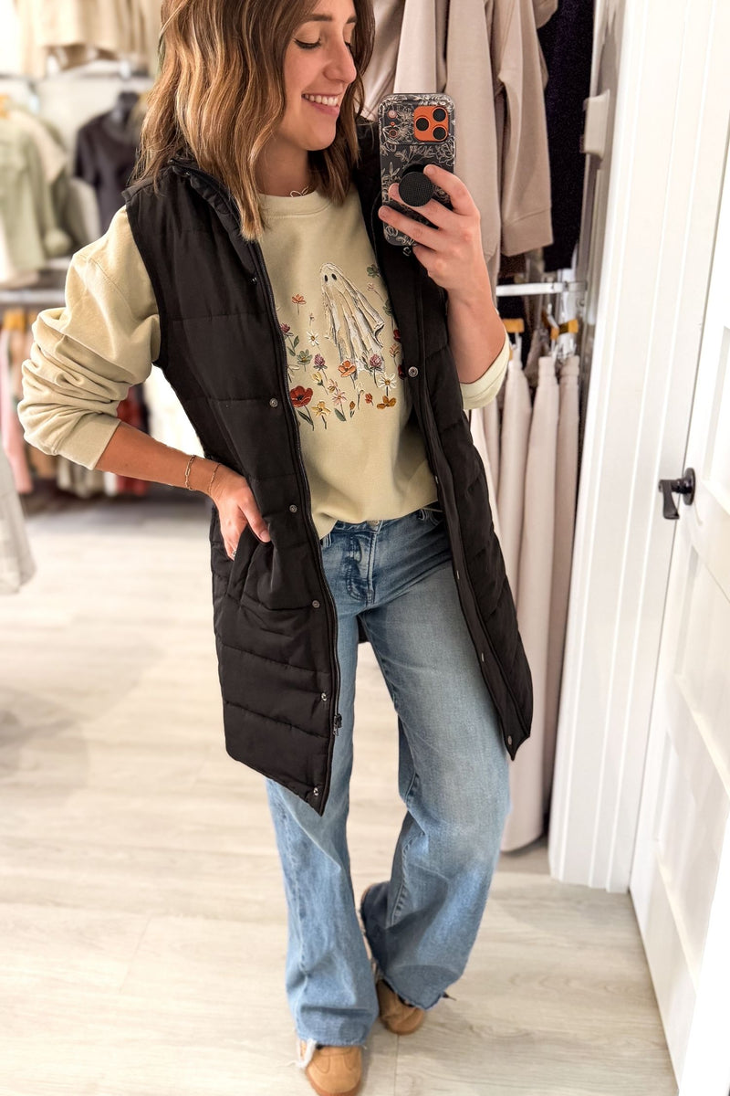 A woman takes a mirror selfie in a clothing boutique wearing a black sleeveless long puffer vest over a beige graphic t-shirt, blue jeans, and tan boots.