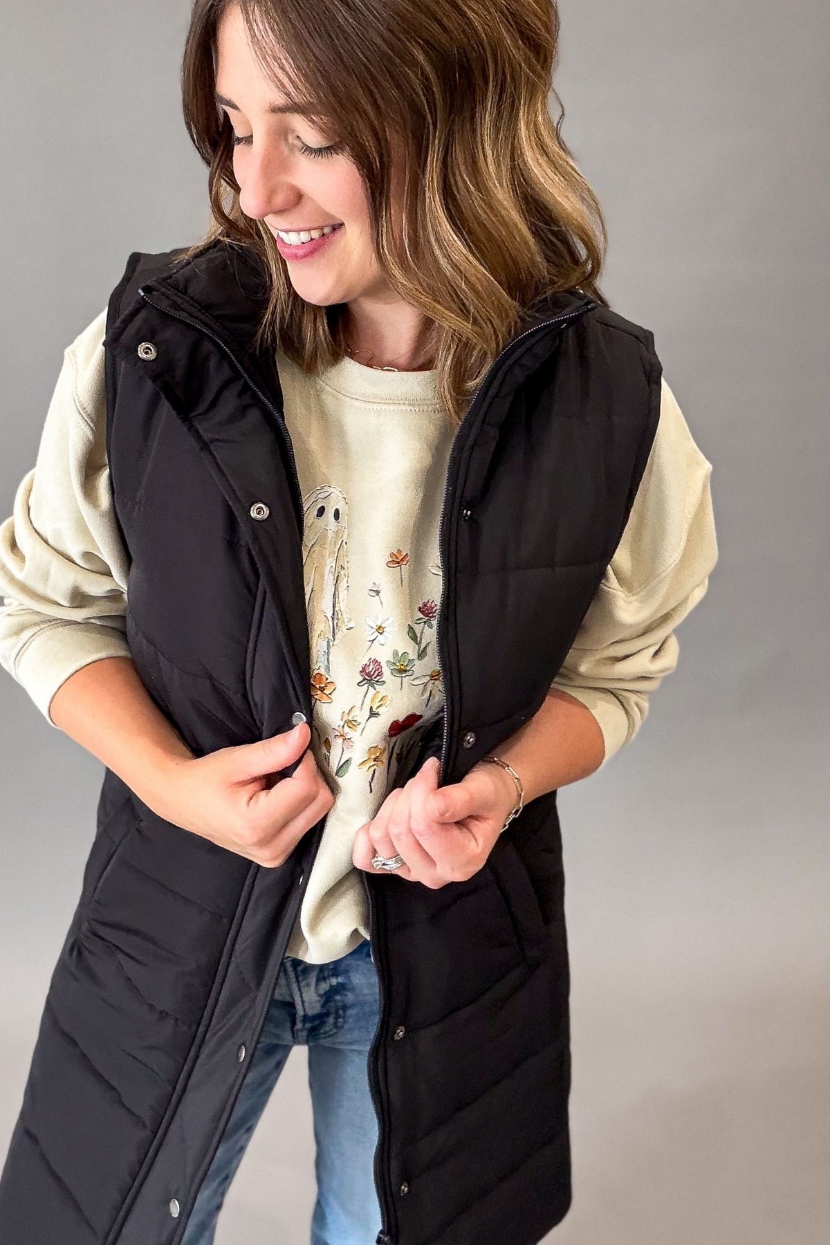A woman wearing a long black puffer vest over a cream sweatshirt and blue jeans, standing against a plain gray studio background.