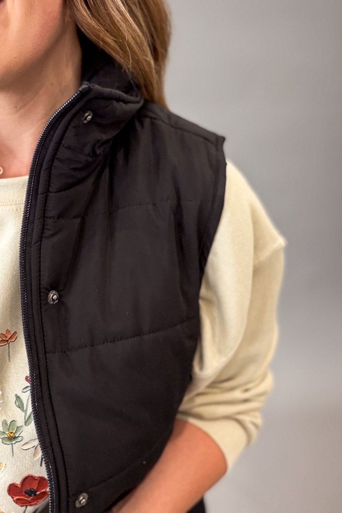 A close-up of a person wearing a black sleeveless puffer vest over a cream-colored long-sleeve top with a floral print.