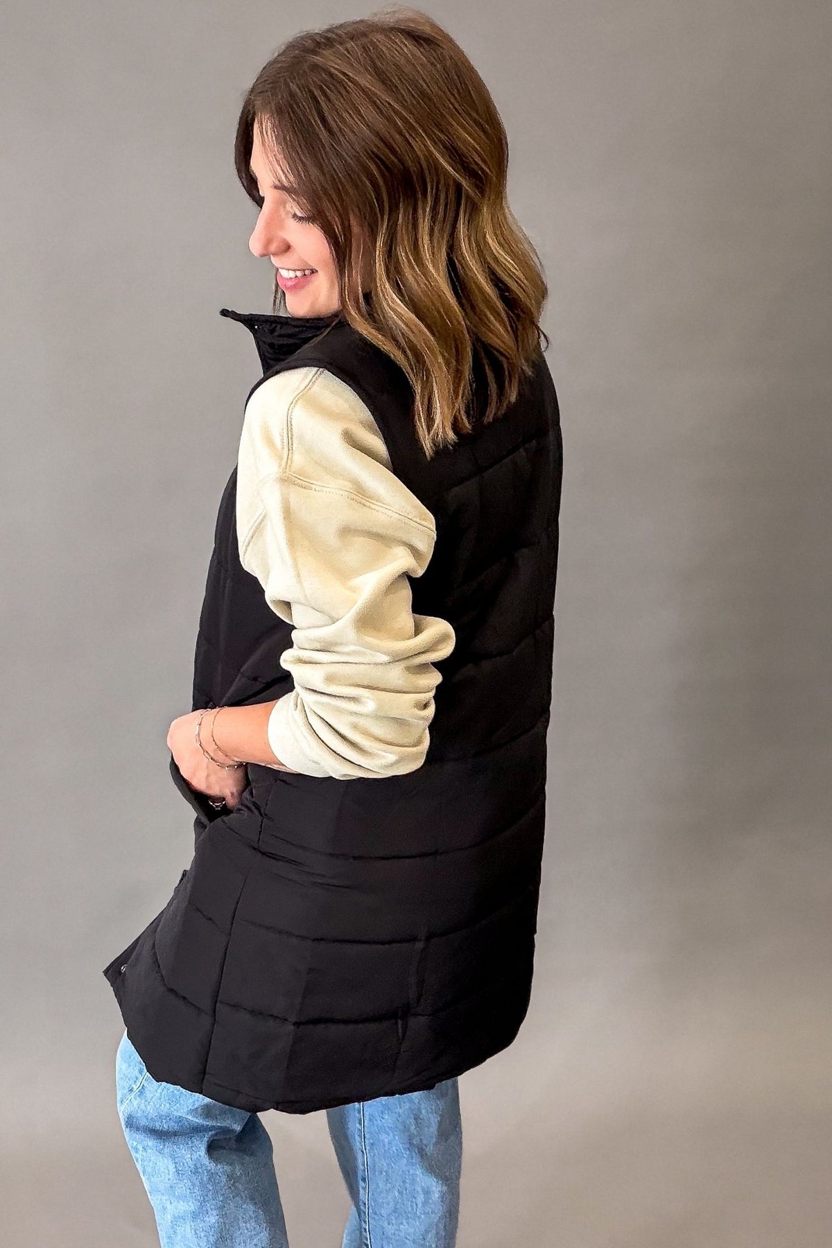 A woman wearing a black sleeveless puffer vest over a cream-colored long-sleeve top and blue jeans, photographed against a neutral gray studio background.