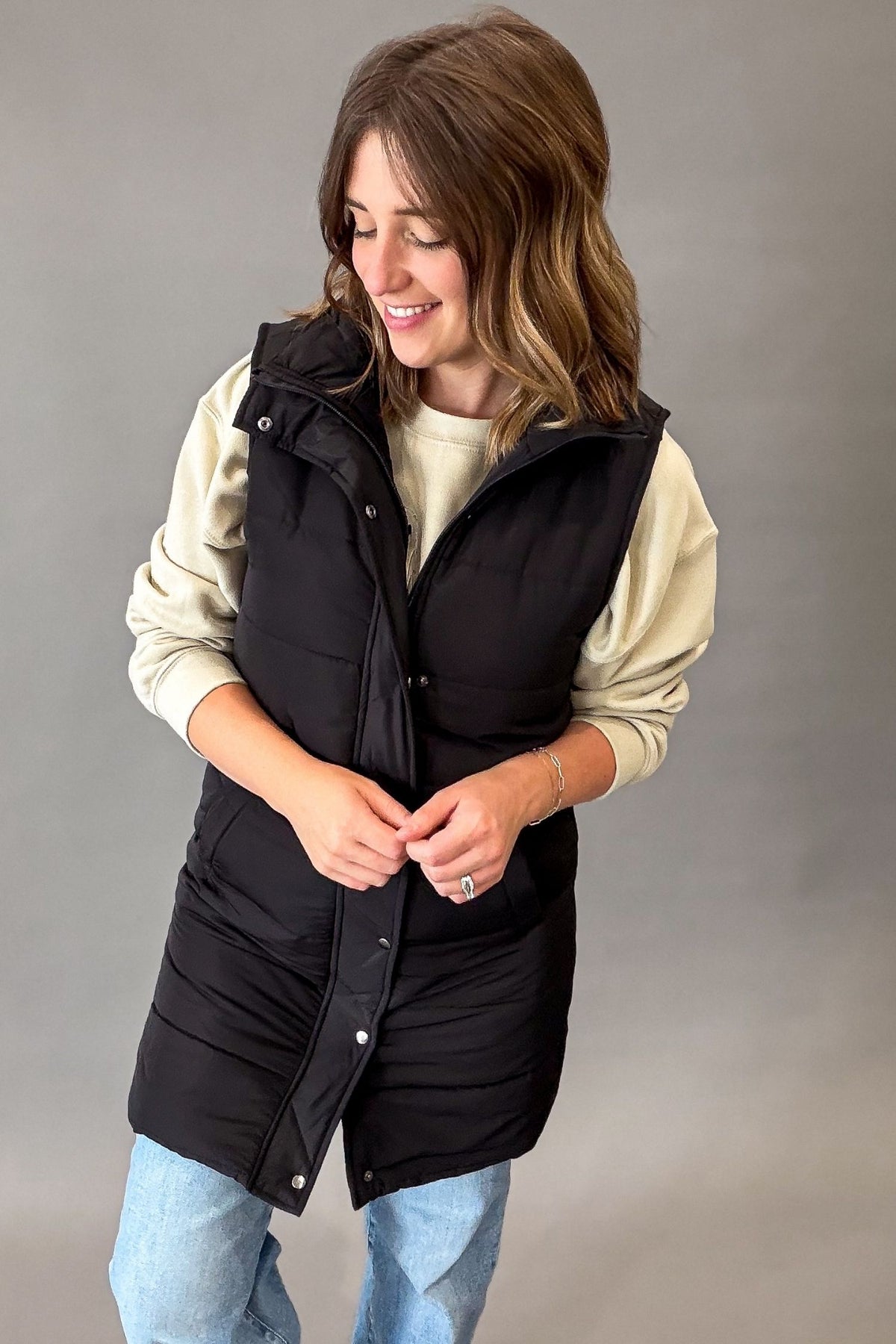 A woman wearing a black sleeveless puffer vest over a cream long-sleeve shirt and light blue jeans, standing against a neutral gray studio background.