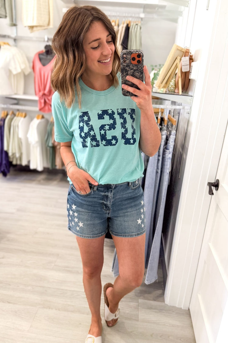 woman taking a mirror selfie wearing a baby blue USA graphic tee paired with judy blue star shorts and white sandals