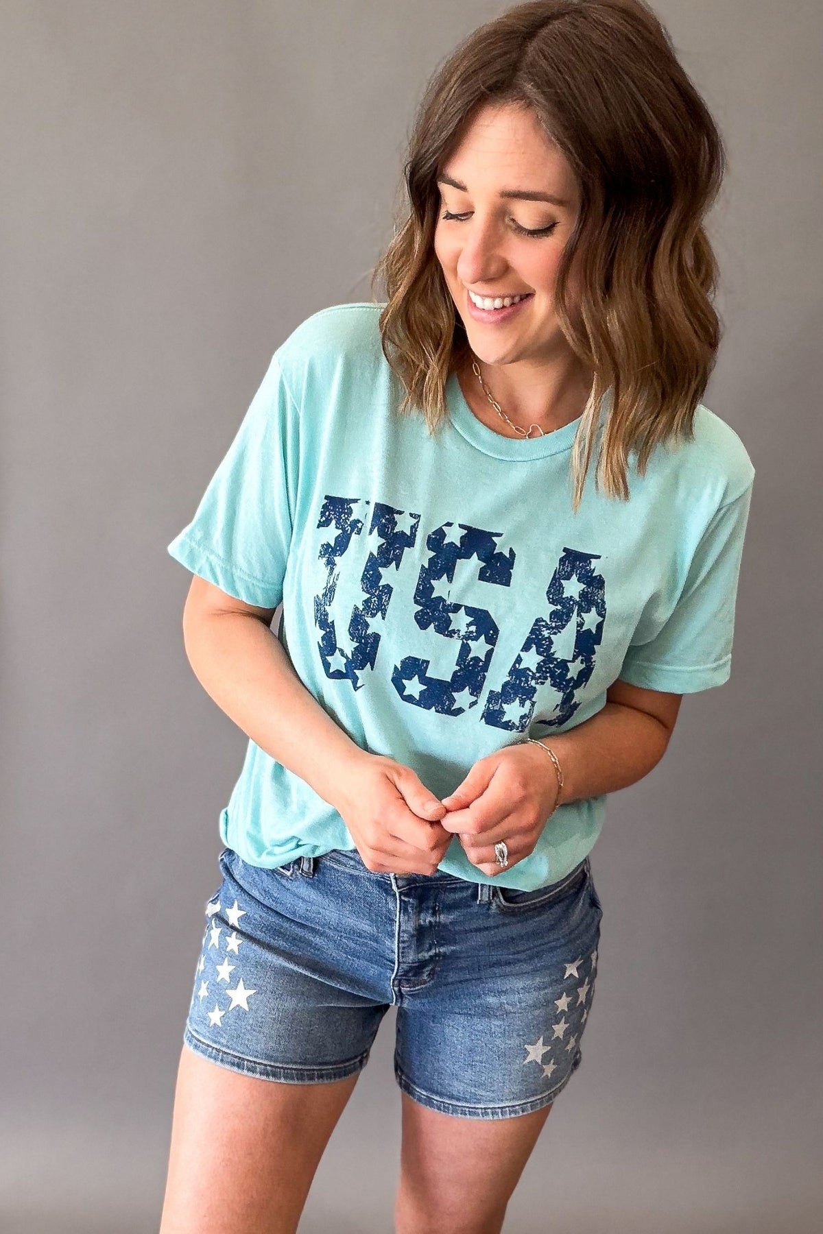 Woman wearing a light blue t-shirt with 'USA' printed in navy and star-patterned denim shorts on a gray background