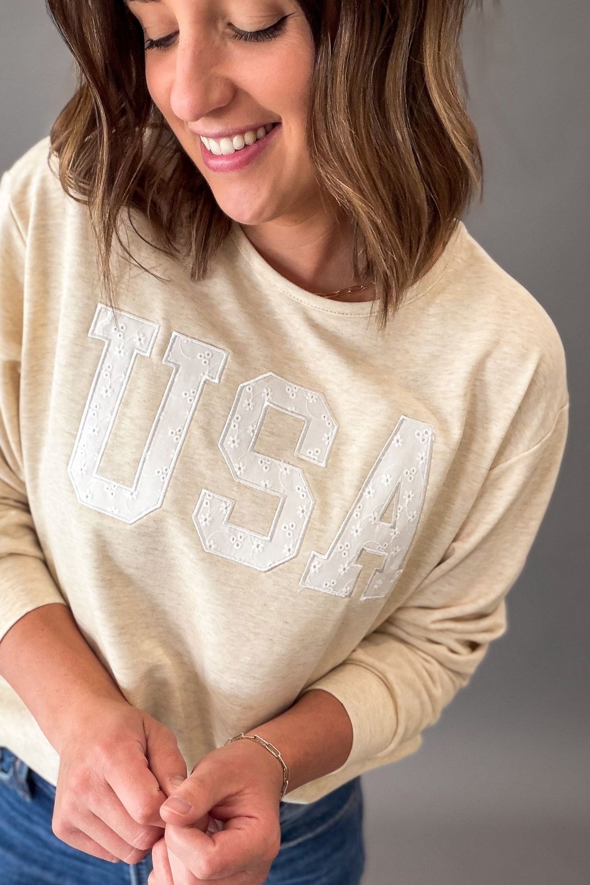 Women's heathered warm taupe USA sweatshirt featuring white eyelet appliqué lettering, crew neckline, drop shoulders, and a relaxed fit paired with denim.