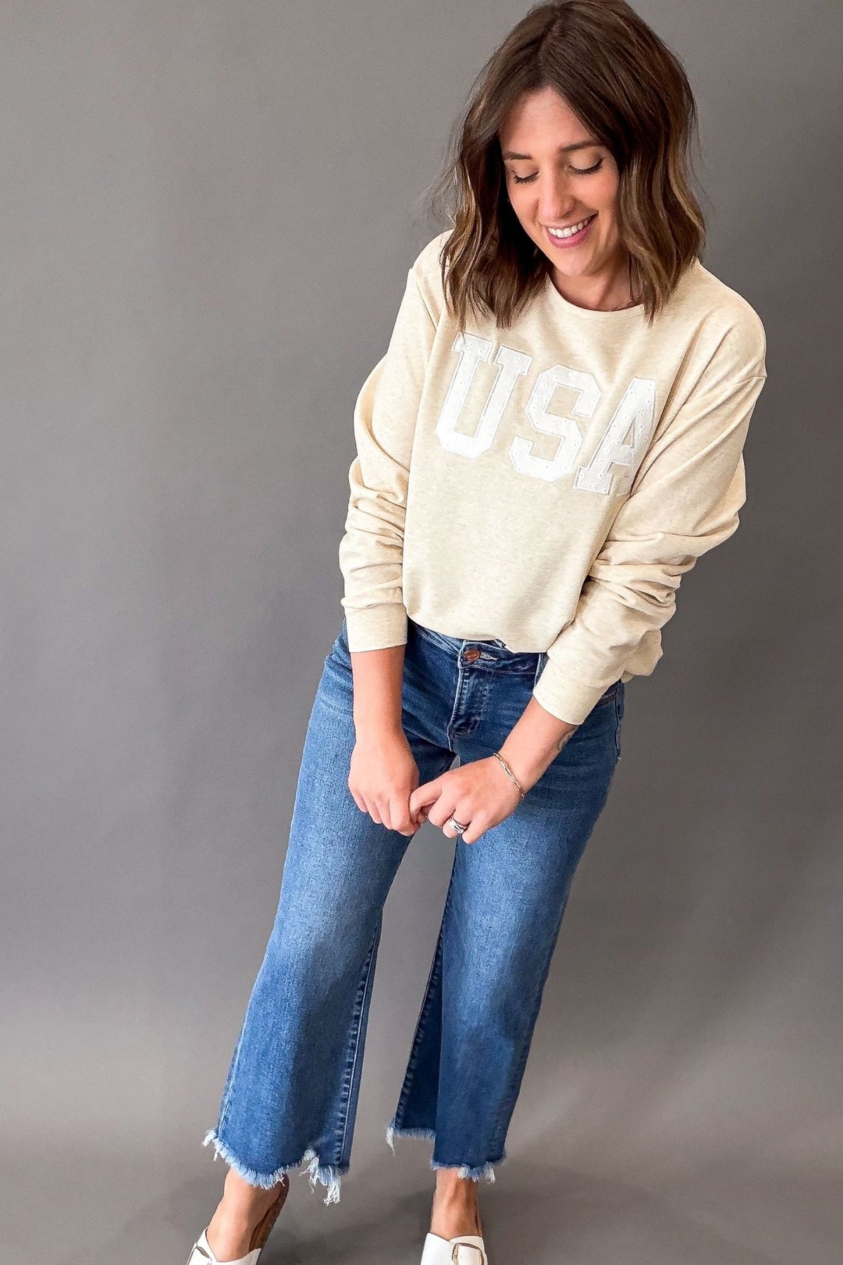 Women's heathered warm taupe USA sweatshirt featuring white eyelet appliqué lettering, crew neckline, drop shoulders, and a relaxed fit paired with denim.