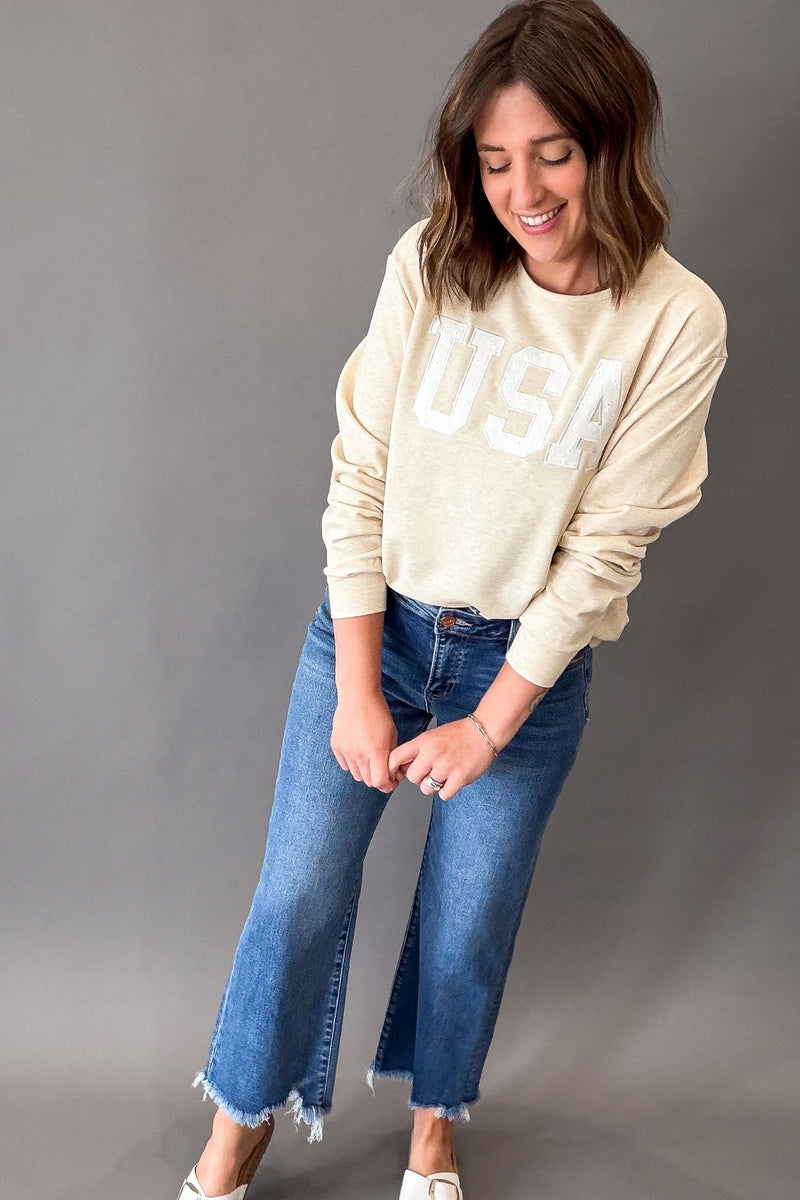 Women's heathered warm taupe USA sweatshirt featuring white eyelet appliqué lettering, crew neckline, drop shoulders, and a relaxed fit paired with denim.