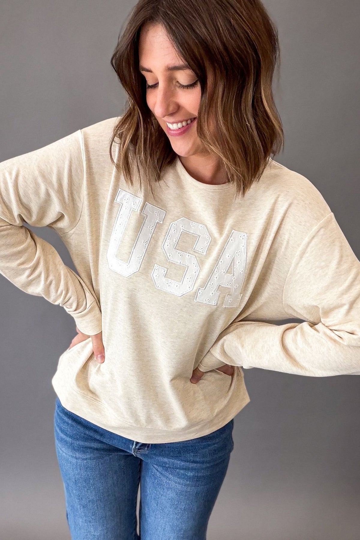 Women's heathered warm taupe USA sweatshirt featuring white eyelet appliqué lettering, crew neckline, drop shoulders, and a relaxed fit paired with denim.