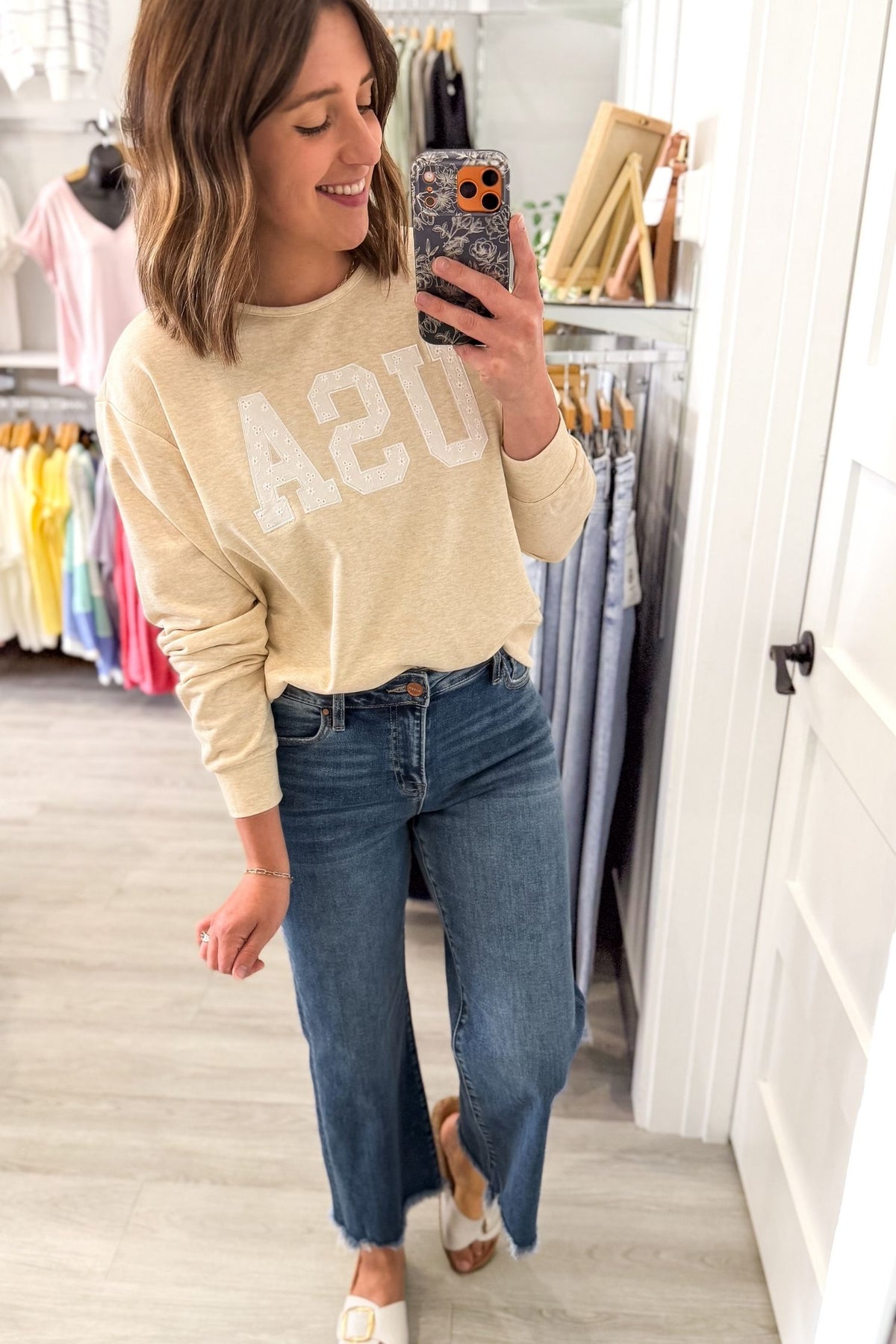 Woman taking a mirror selfie wearing a beige sweatshirt with 'USA' text and blue jeans in a clothing store.