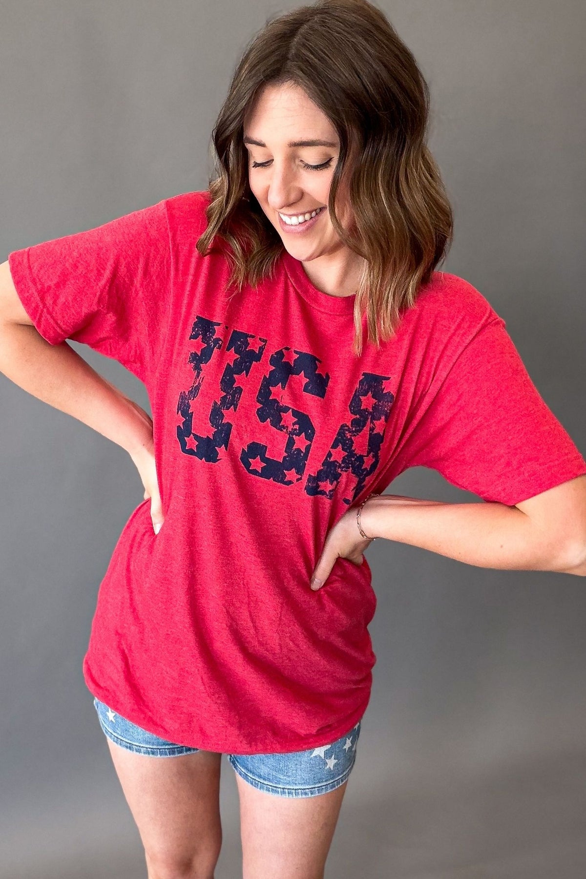 woman wearing a usa graphic tee untucked to show length with judy blue shorts on a grey background