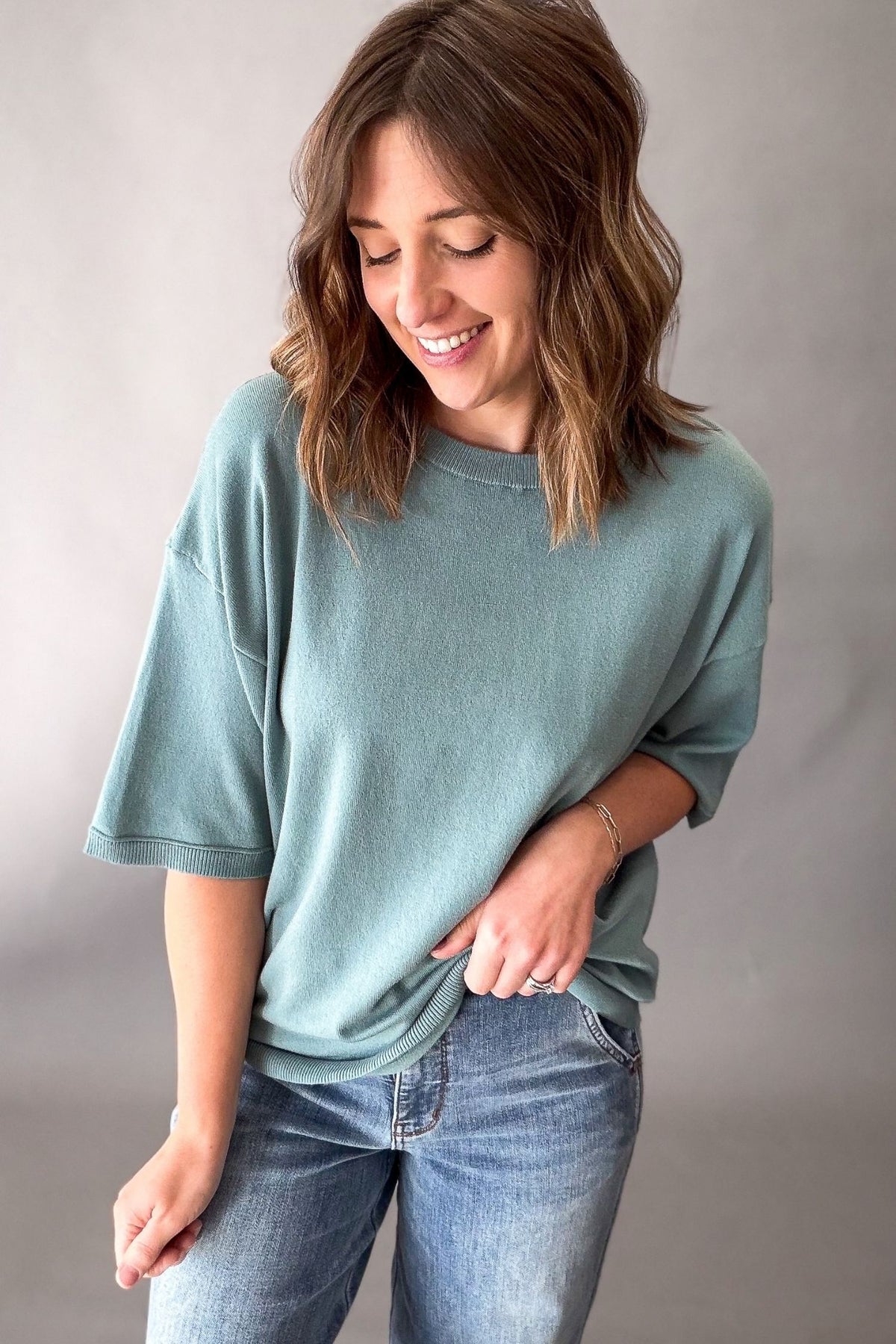 A woman wearing a seafoam colored oversized short-sleeve t-shirt and blue jeans, standing against a neutral gray studio background.
