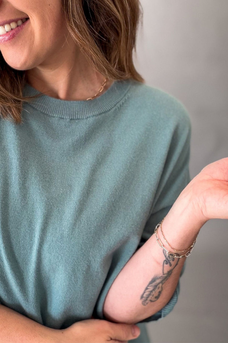 A close-up lifestyle shot of a woman wearing a soft seafoam crewneck sweater, showing her arm with a feather tattoo and delicate gold bracelets.
