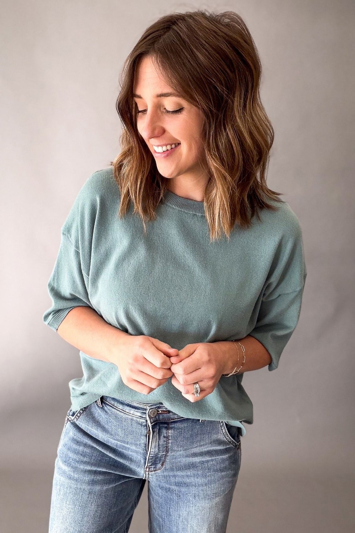 A woman with shoulder-length brown hair wearing a seafoam crewneck sweatshirt and blue jeans, standing against a neutral gray studio background.