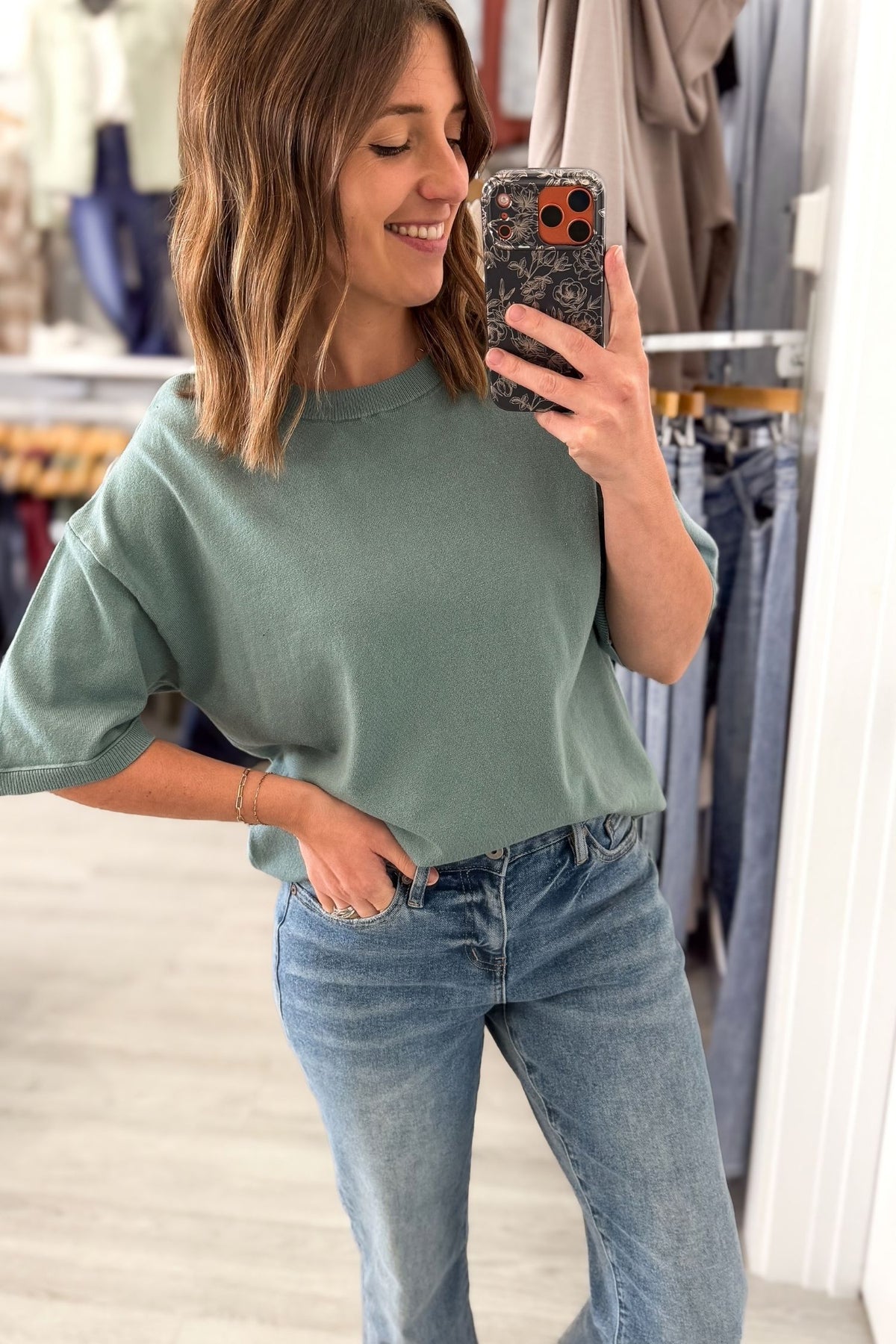 A woman takes a mirror selfie in a clothing store, wearing an oversized seafoam crewneck sweatshirt and light-wash blue jeans.