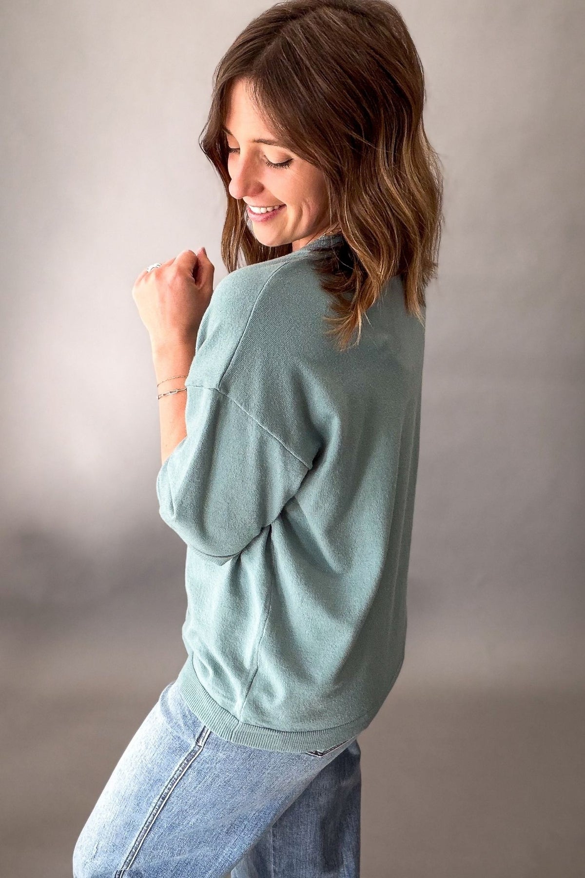A woman wearing a seafoam long-sleeve crewneck sweatshirt and light blue jeans, shown from a side-back angle against a neutral gray studio background.