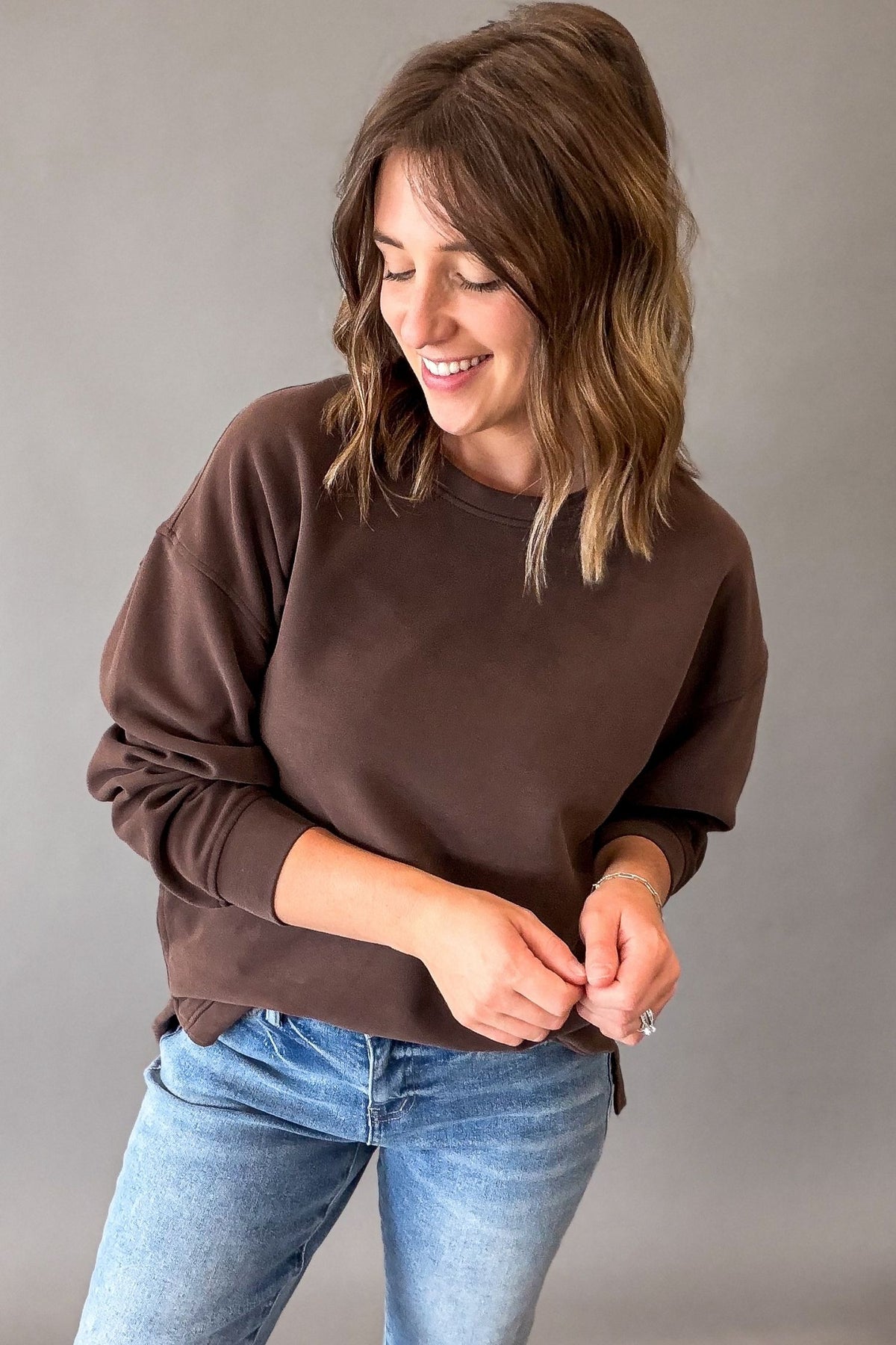 A woman wearing a brown crewneck sweatshirt and light blue jeans, standing against a neutral gray studio background.