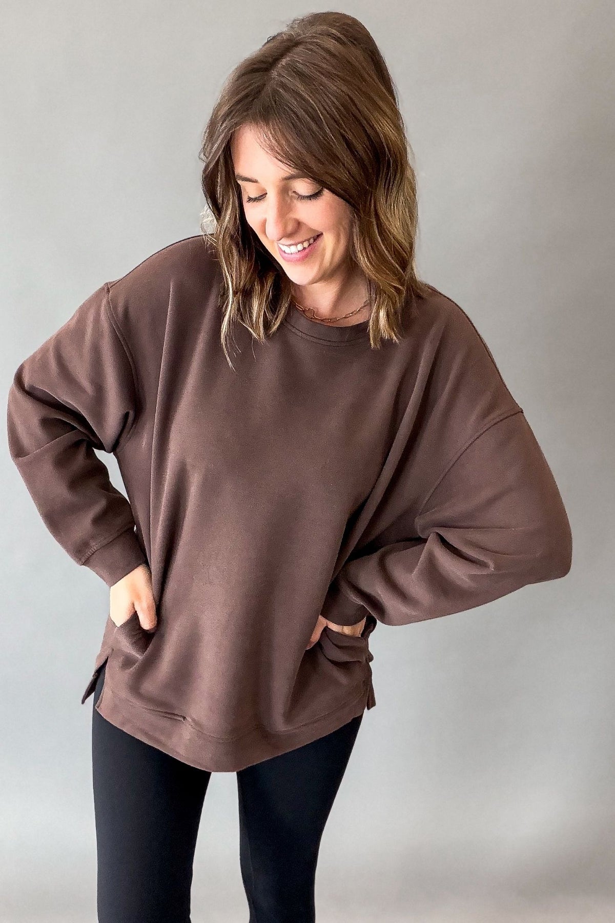A woman wearing an oversized brown crewneck sweatshirt and black leggings, standing against a neutral gray studio background.