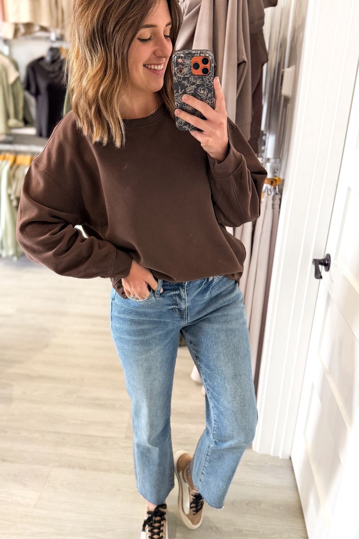 A woman takes a mirror selfie in a clothing store, wearing a brown crewneck sweatshirt and light-wash blue jeans.