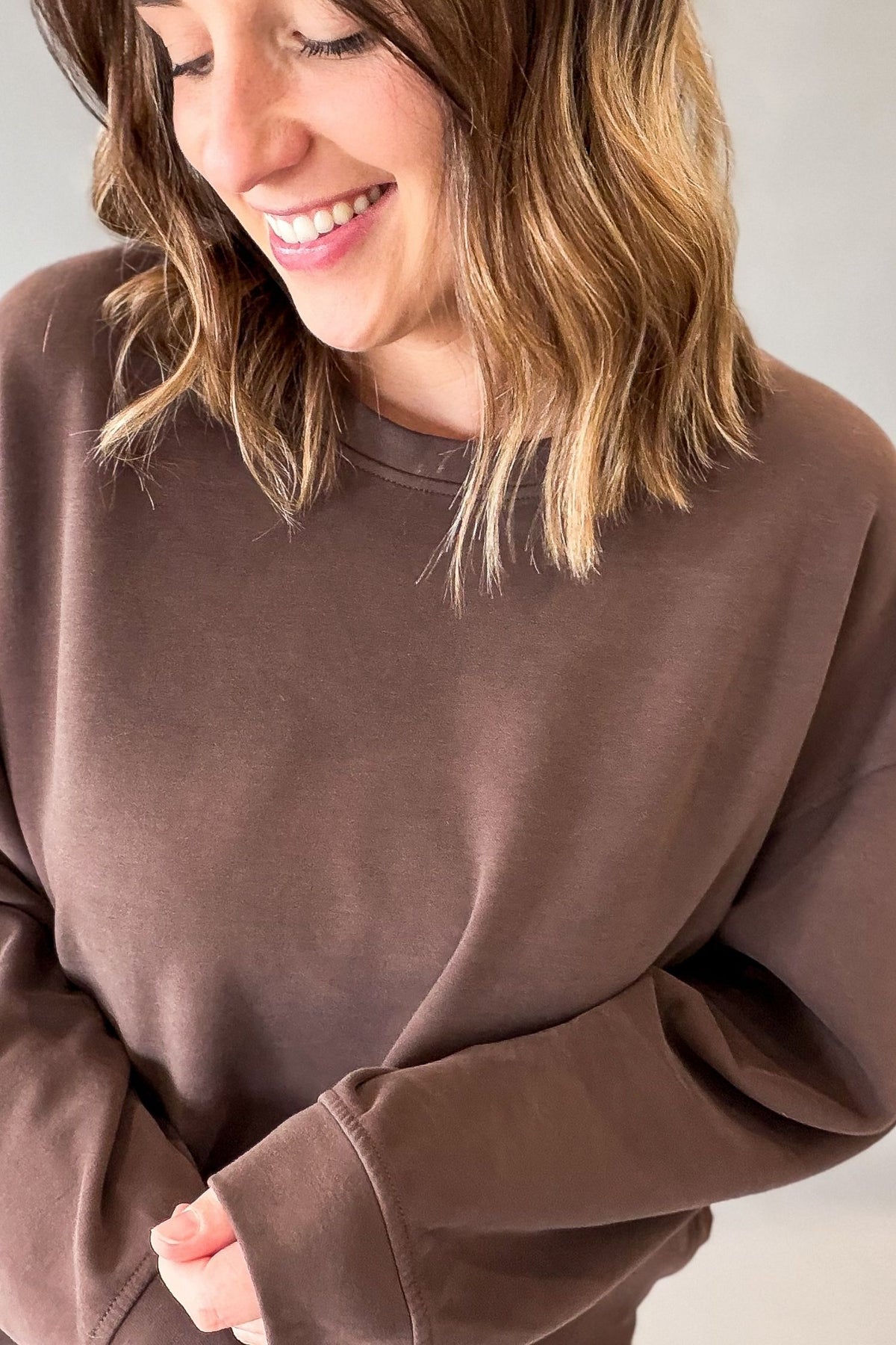 A woman wearing a brown crewneck sweatshirt, shown from the chest up against a plain light gray background.