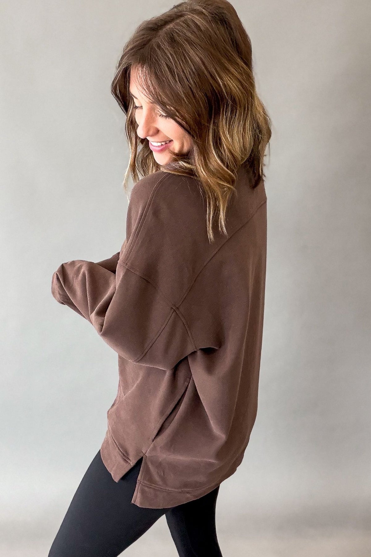 A woman wearing an oversized brown long-sleeve top and black leggings, photographed from the back against a plain light gray studio background.