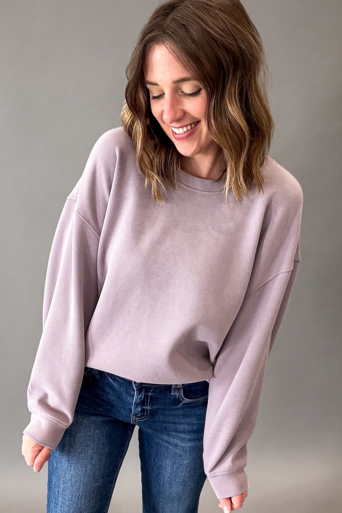 A woman wearing a dusty lavender crewneck sweatshirt and blue jeans, smiling and looking down against a solid gray studio background.