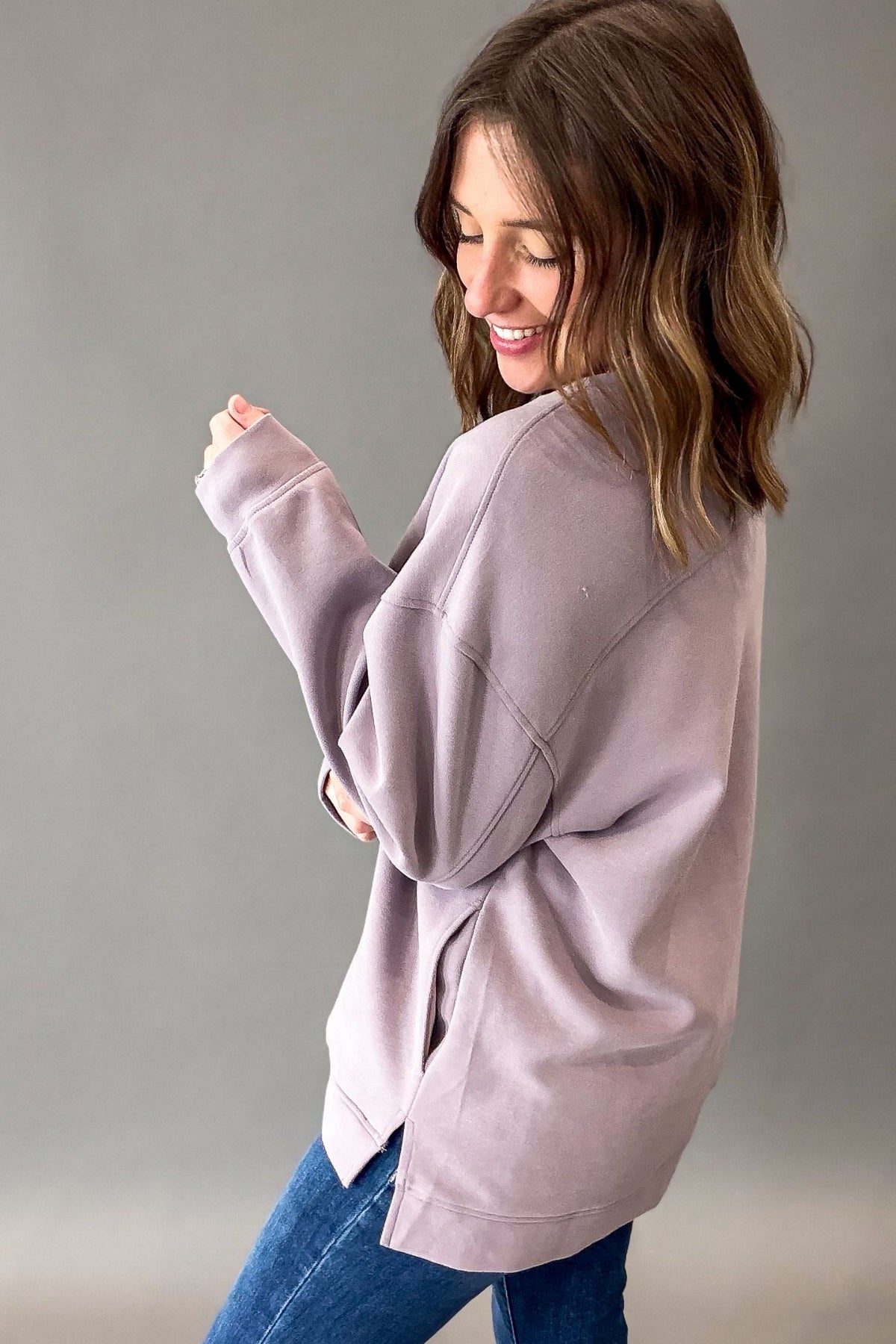 A woman wearing an oversized dusty lavender sweatshirt and blue jeans, shown from a side-back angle against a plain gray studio background.