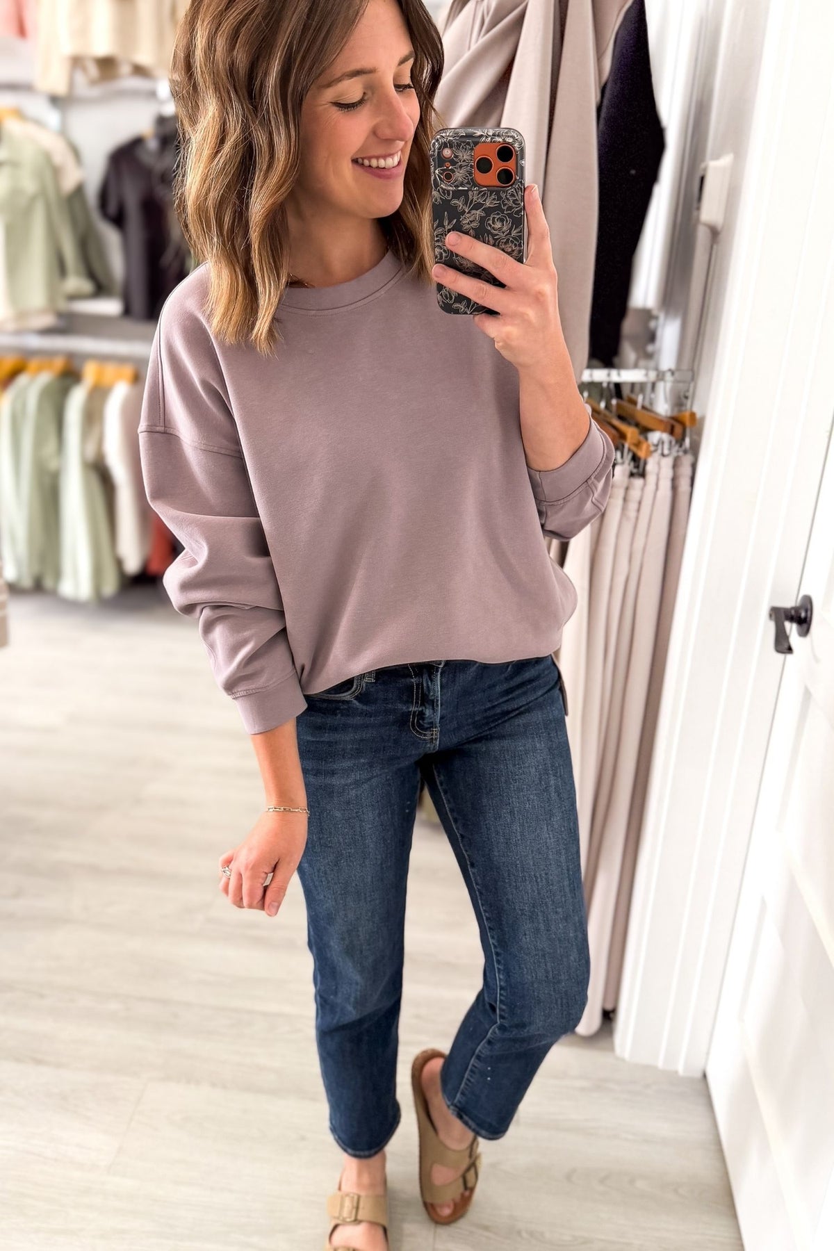 A woman takes a mirror selfie in a clothing boutique, wearing a dusty lilac crewneck sweatshirt and dark wash blue jeans.