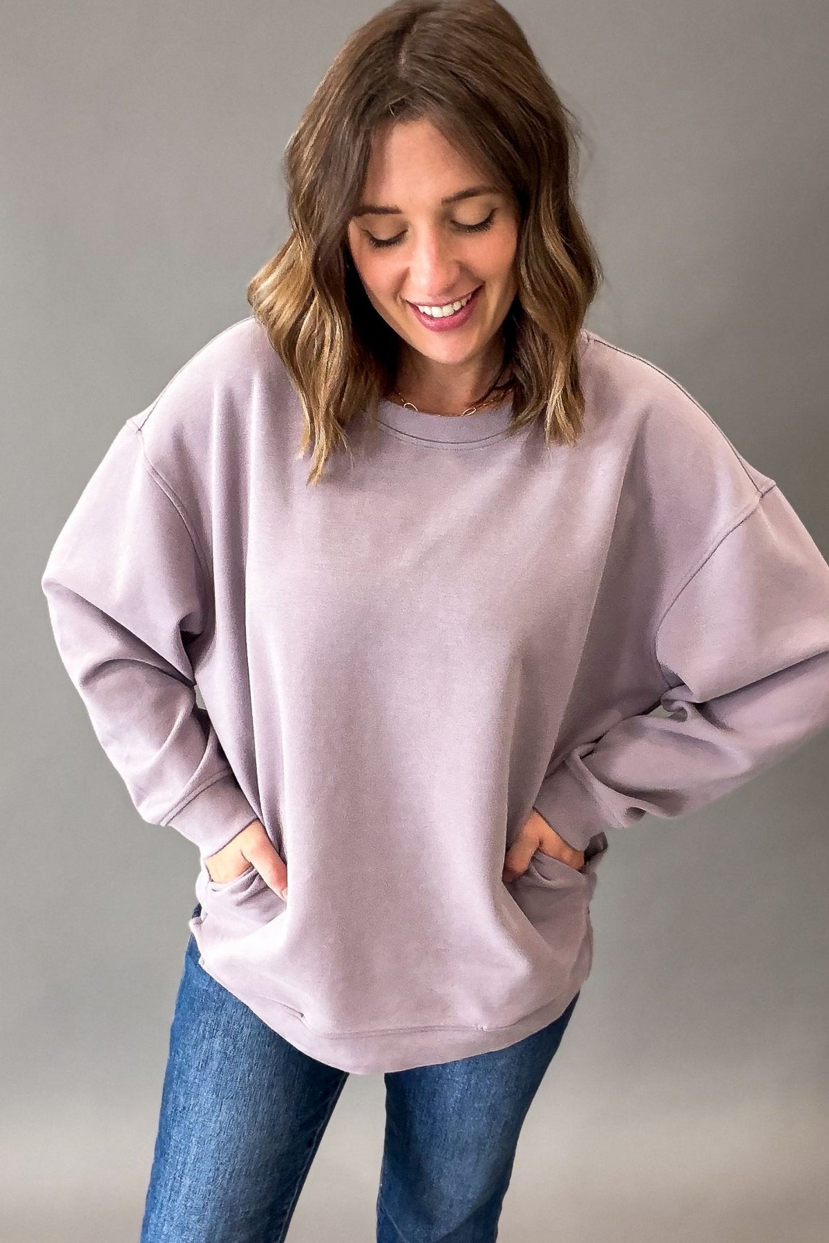 A woman wearing an oversized dusty lavender crewneck sweatshirt and blue jeans, standing against a neutral gray studio background.