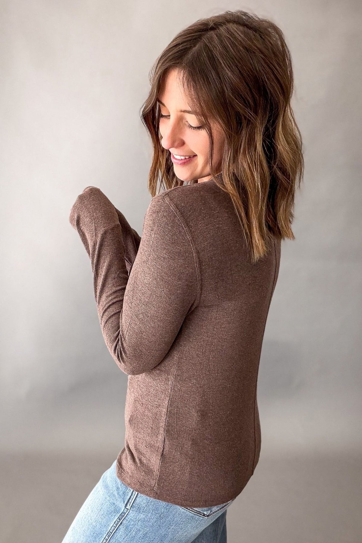 A woman with shoulder-length brown hair is shown from the back wearing a fitted, long-sleeve knit top in a muted taupe-brown color, paired with light blue jeans, against a neutral gray studio background.