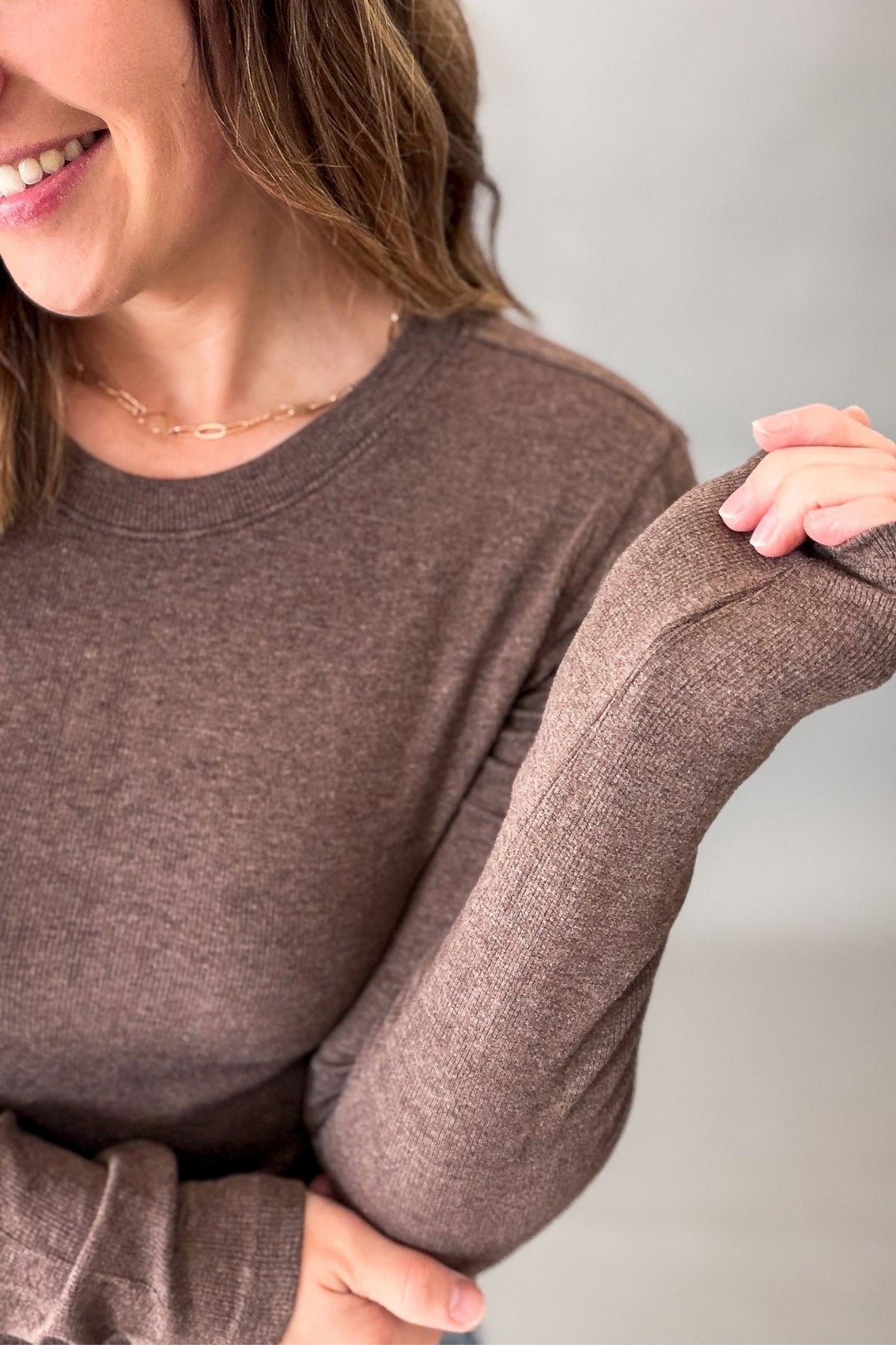A close-up of a woman wearing a heathered brown crewneck sweater, showing the texture of the fabric and the fit of the long sleeves.
