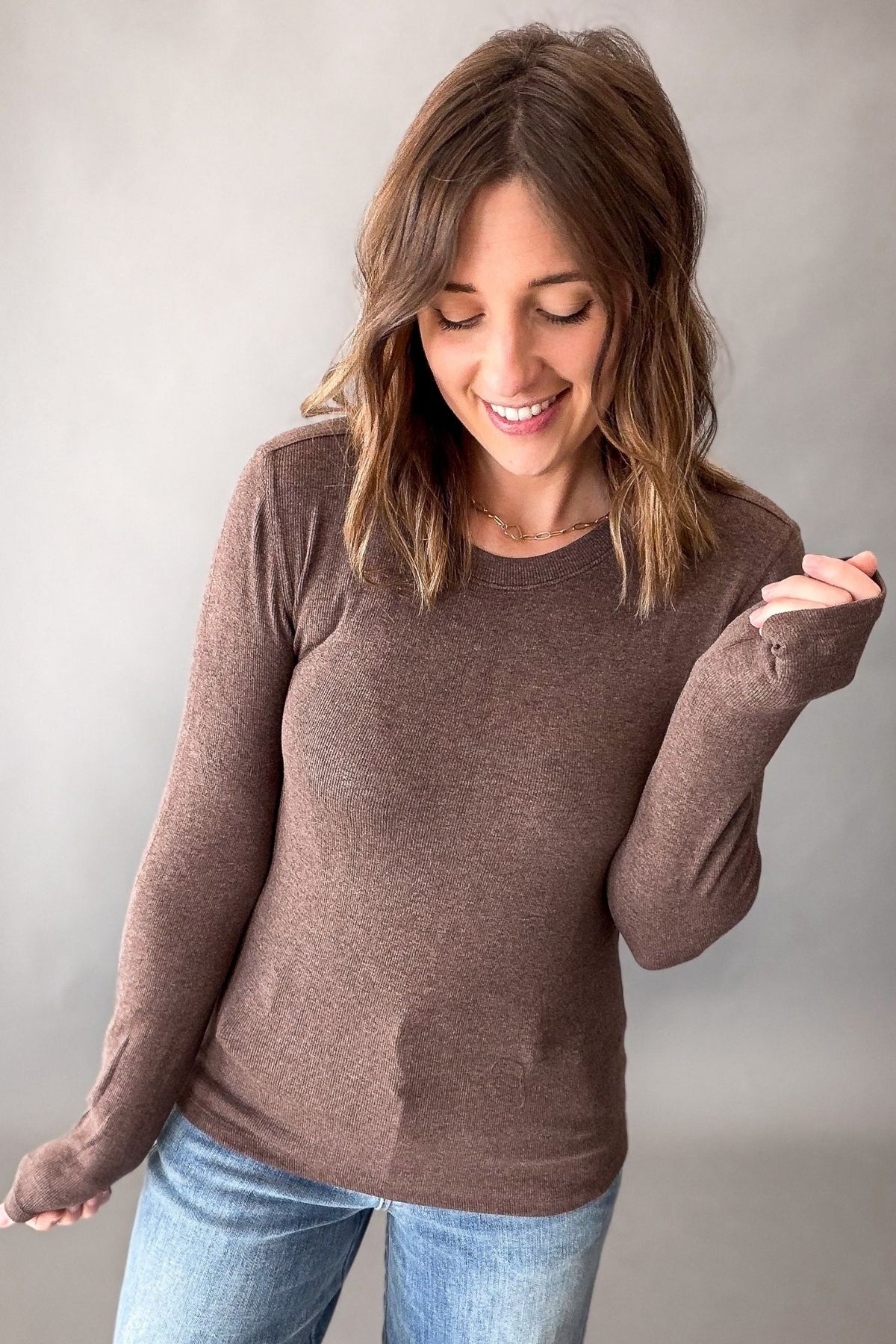 A woman wearing a heathered brown long-sleeve knit top with thumbholes and blue jeans, posing against a neutral gray studio background.
