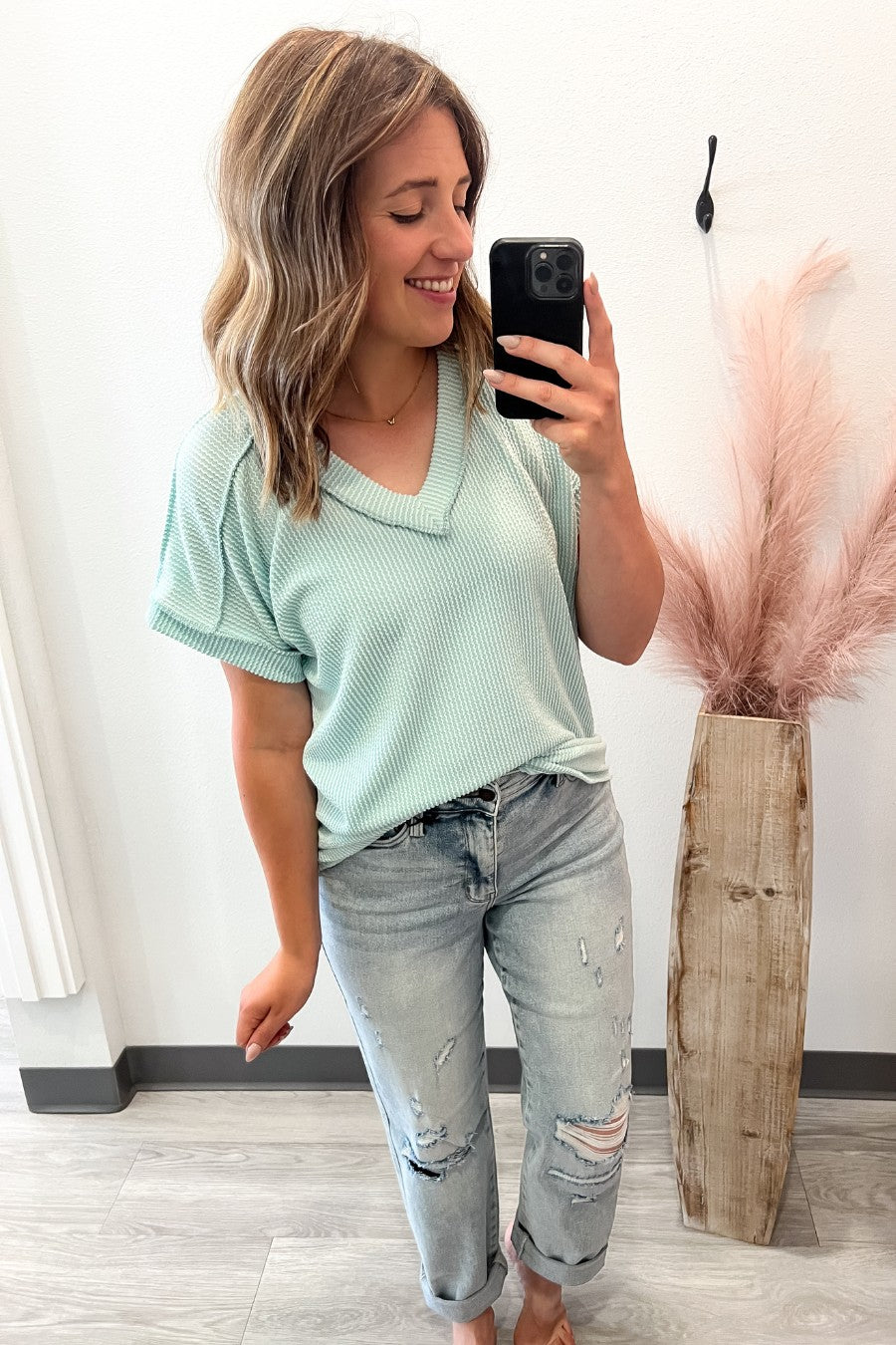 Woman taking a selfie in a light mint top and ripped jeans, standing next to a vase with pink decorative elements.