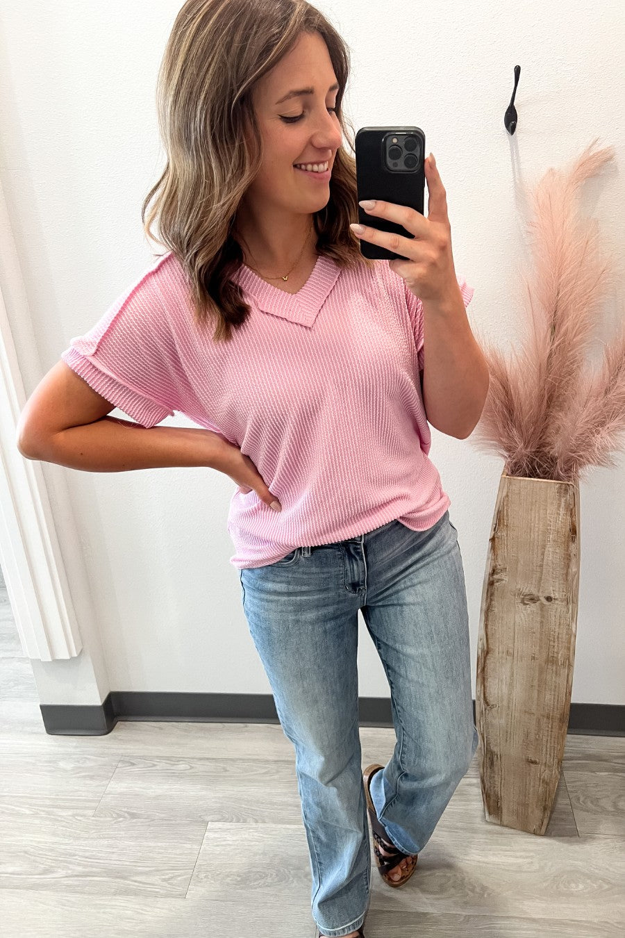 Woman taking a mirror selfie wearing a pink ribbed shirt and blue jeans, standing next to a vase with dried plants.