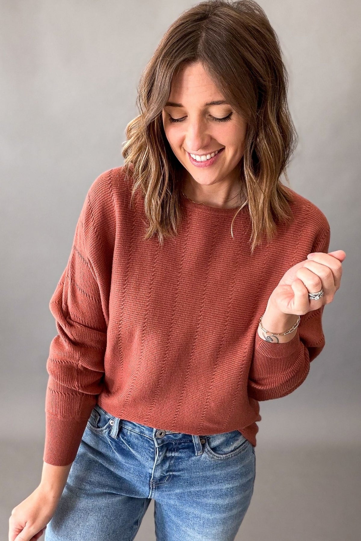 A woman wearing a textured, muted terracotta-colored knit sweater and blue jeans, standing against a neutral gray studio background.