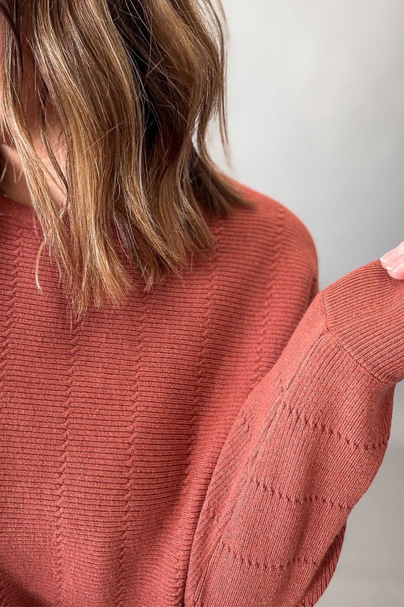 Close-up of a person wearing a textured knit sweater in a muted terracotta or dusty rose color, with shoulder-length brown hair visible against a plain light background.
