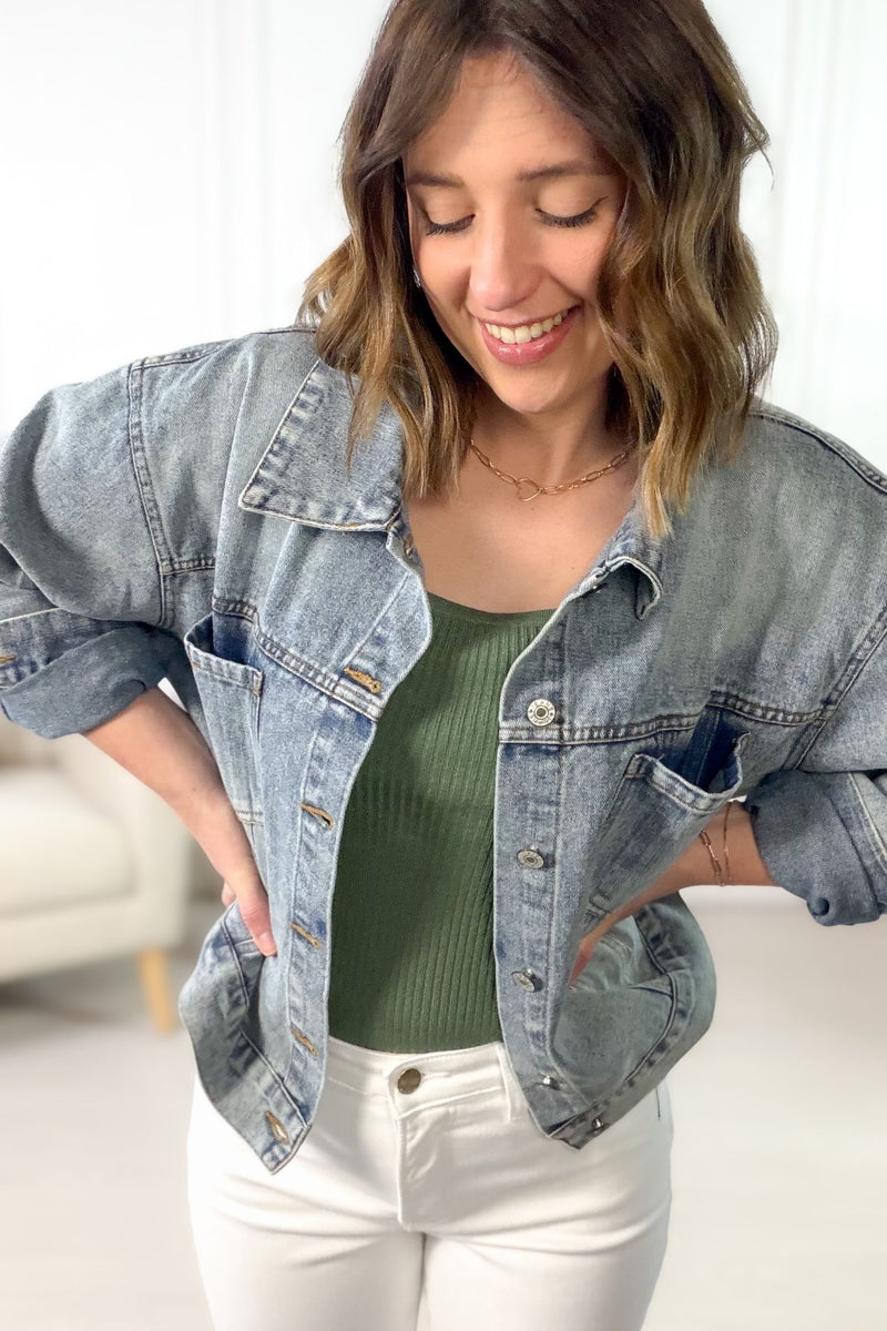 Woman wearing a denim jacket over a green top and white pants, smiling.