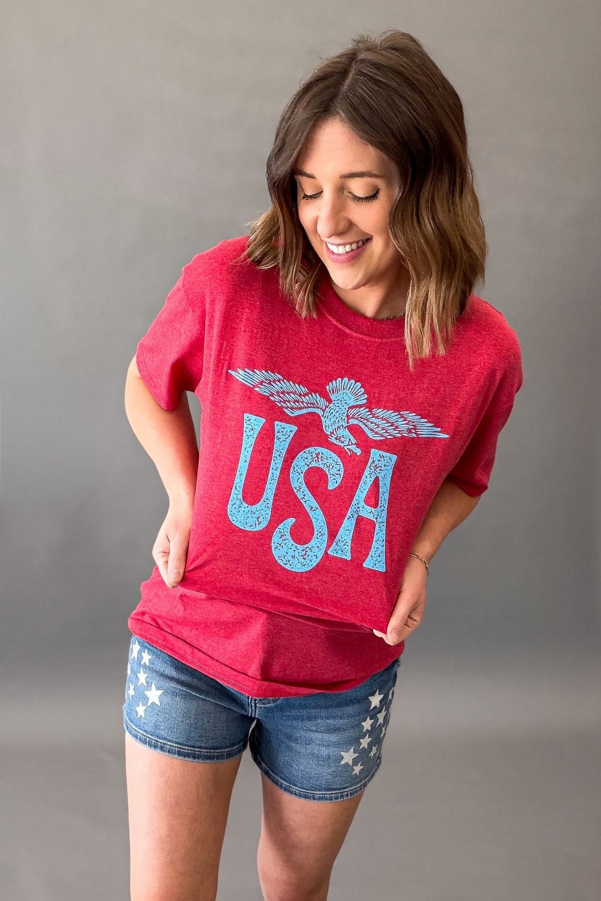 Woman wearing a red vintage USA eagle graphic tee with distressed light blue screen printed graphic and denim shorts showing the distressed details on the graphic