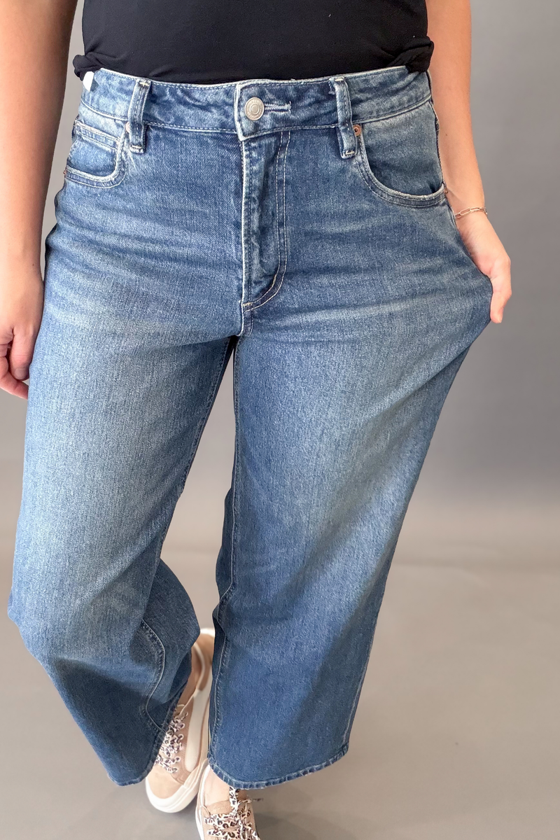 A person wearing a pair of wide-leg blue denim jeans with a faded wash, paired with a black top and patterned sneakers, shown from the waist down against a neutral gray studio background.