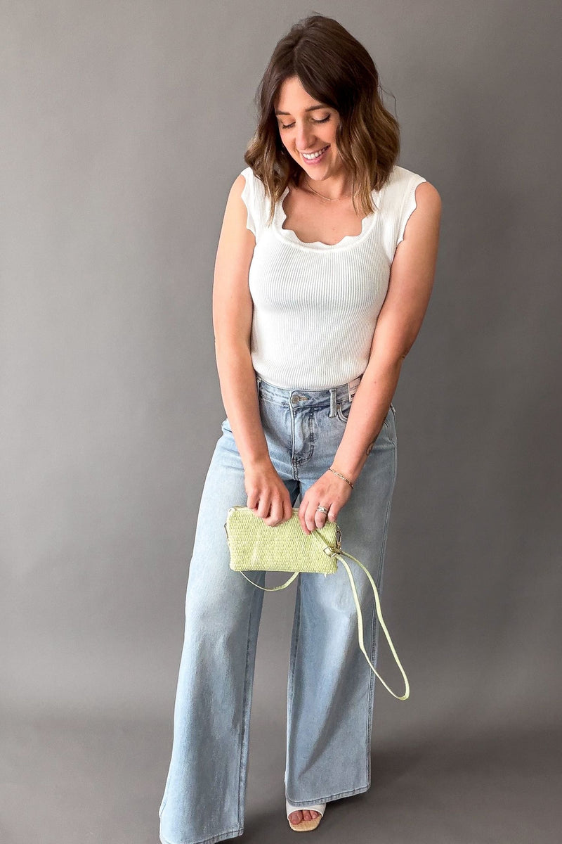 Woman holding a green handbag against a gray background wearing a white ribbed scalloped edge tank and blue jeans