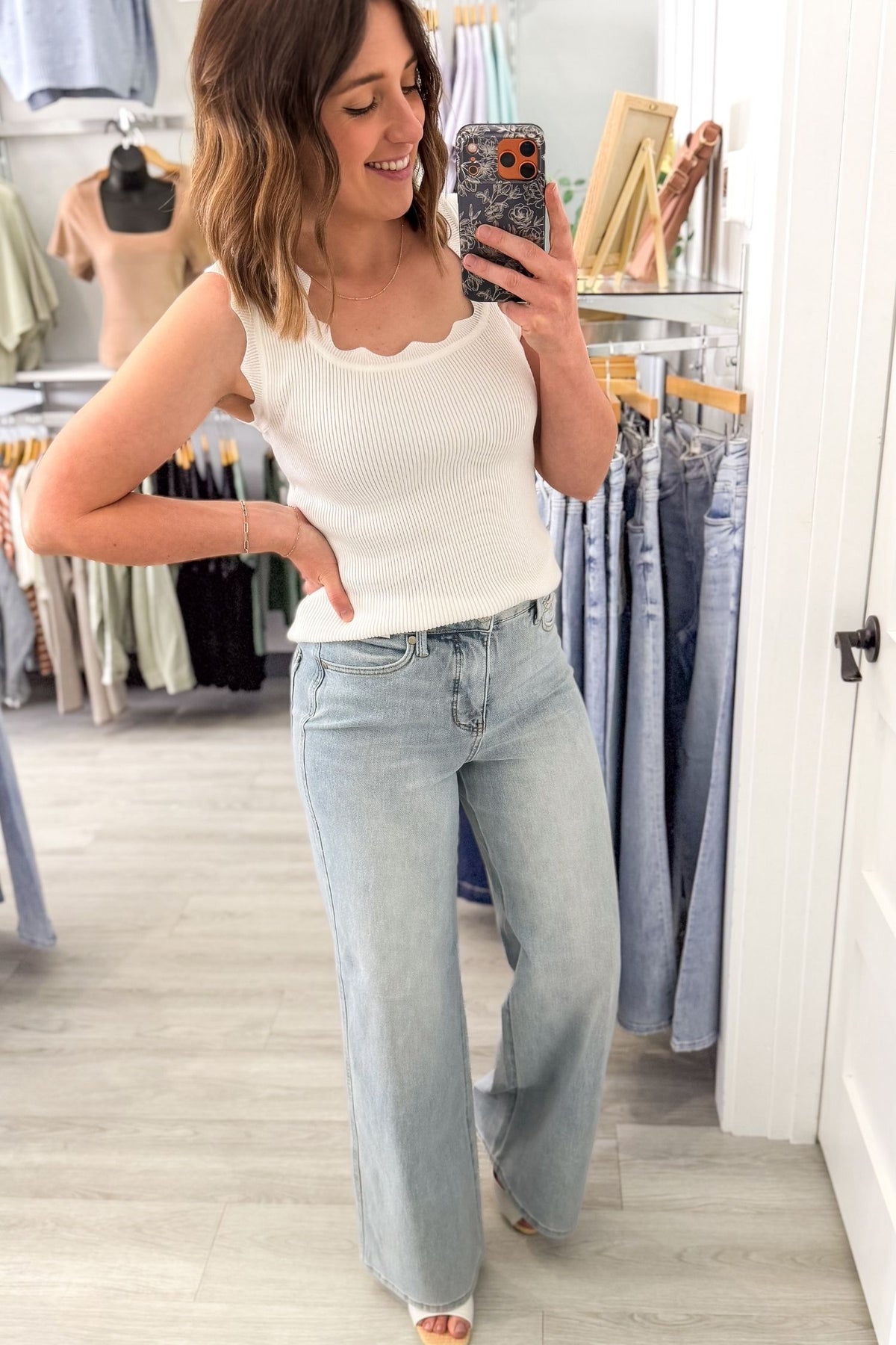 Woman taking a mirror selfie wearing a white tank top and light blue jeans in a clothing store.