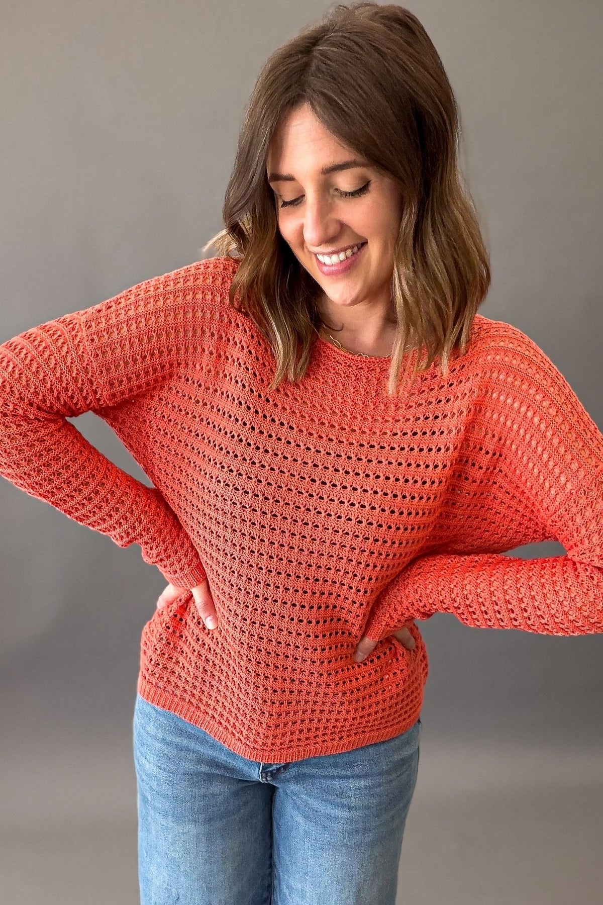 woman wearing an orange crochet pullover long sleeve with blue jeans on a charcoal background