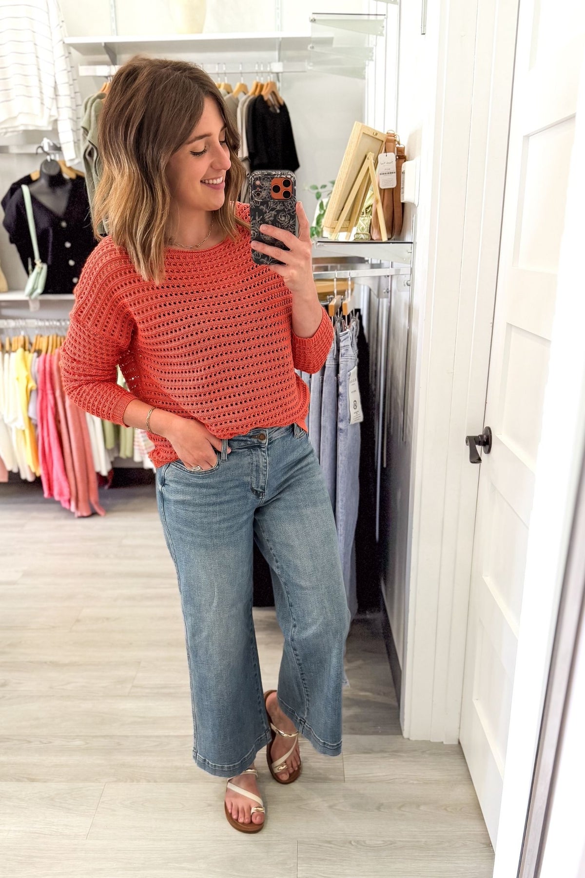 woman taking a mirror selfie wearing an orange wide neck crochet sweater with Judy blue jeans and sandals in a boutique