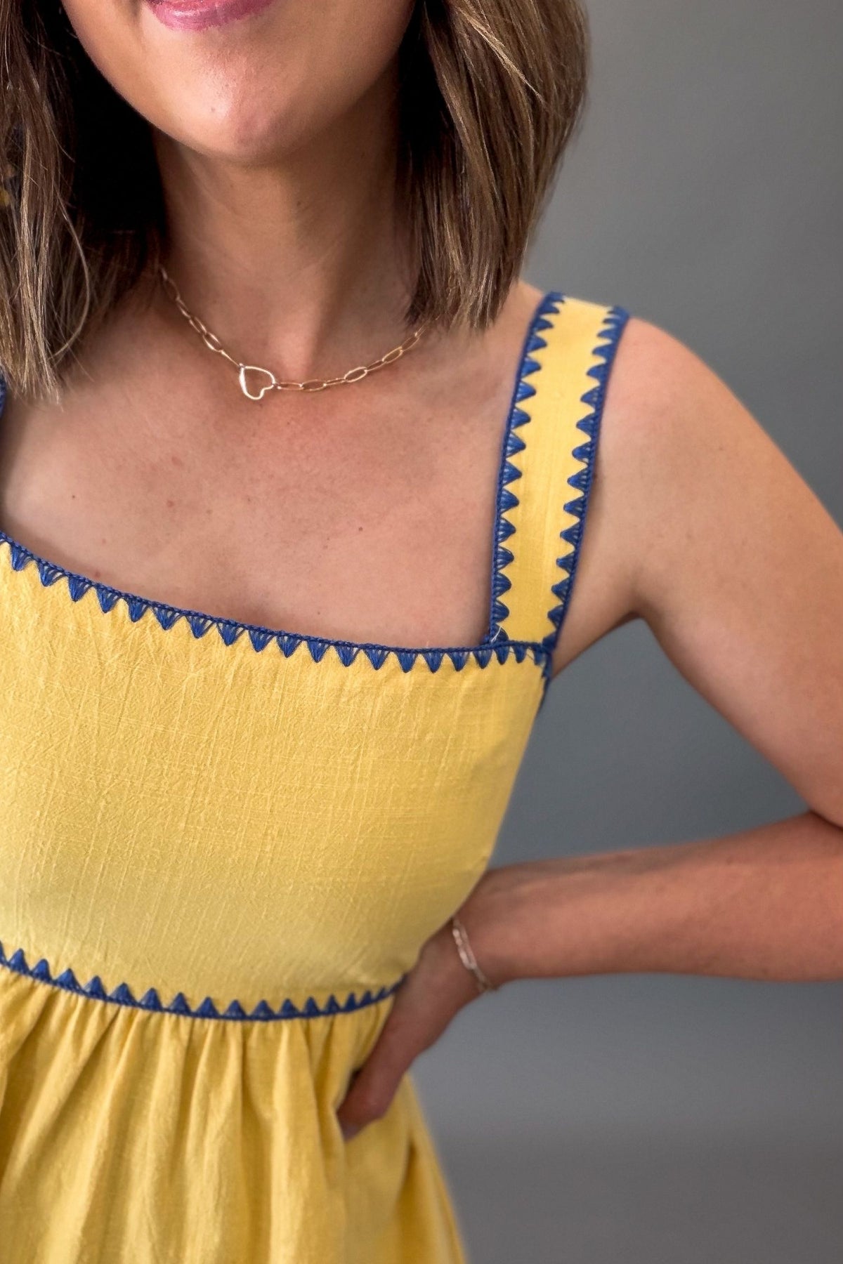 Woman wearing a sunny yellow midi dress featuring a square neckline, blue contrast trim, adjustable shoulder straps, tie-back detail, side pockets, and a flowy lined skirt.