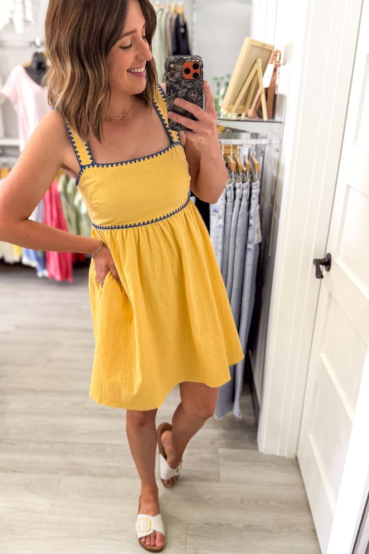 Woman taking a selife wearing a sunny yellow midi dress featuring a square neckline, blue contrast trim, adjustable shoulder straps, tie-back detail, side pockets, and a flowy lined skirt.