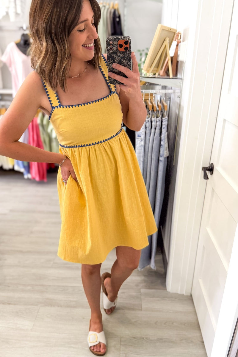 Woman taking a selife wearing a sunny yellow midi dress featuring a square neckline, blue contrast trim, adjustable shoulder straps, tie-back detail, side pockets, and a flowy lined skirt.