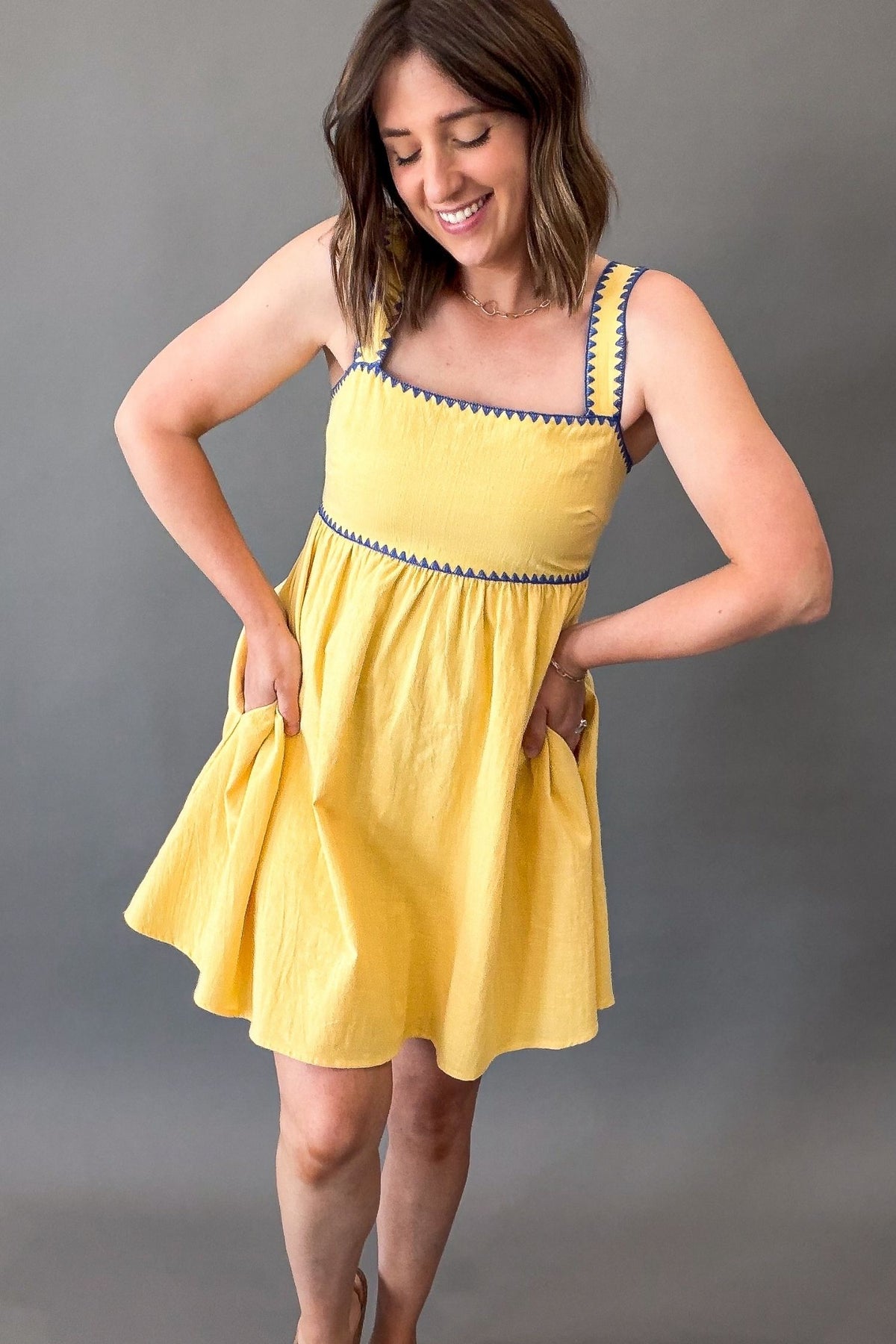 Woman wearing a sunny yellow midi dress featuring a square neckline, blue contrast trim, adjustable shoulder straps, tie-back detail, side pockets, and a flowy lined skirt.